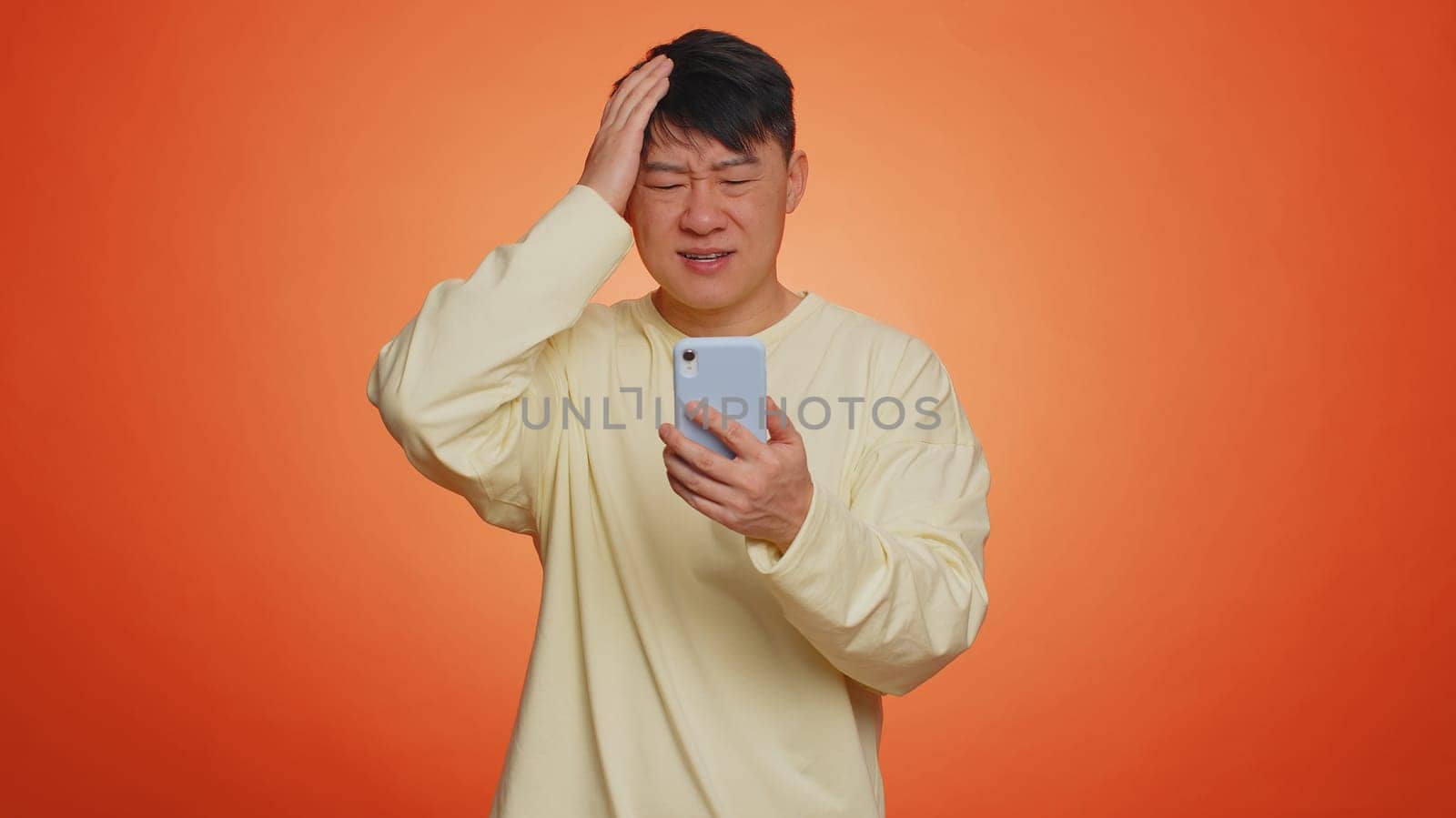 Asian man use mobile cell phone typing browsing, loses becoming surprised sudden lottery results, bad fortune, loss, fail. Handsome chinese guy isolated alone on orange studio wall background, indoors