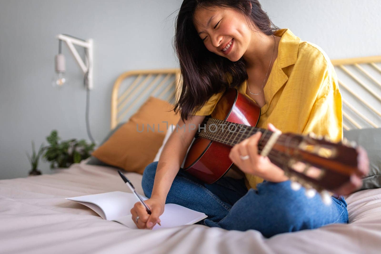 Young Asian female song writer composing song inner bedroom. Writing lyrics. Playing acoustic guitar. Musician by Hoverstock