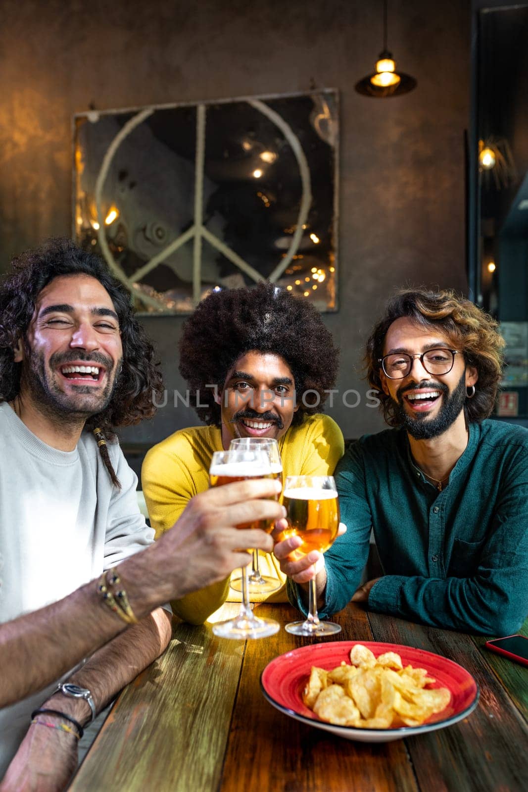 Happy young man showing to multiracial guy friends mobile phone while having drinks together in pub. Technology and leisure activity.
