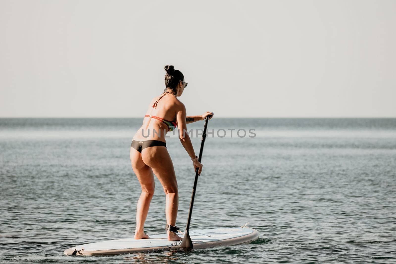 Sea woman sup. Silhouette of happy middle aged woman in rainbow bikini, surfing on SUP board, confident paddling through water surface. Idyllic sunset. Active lifestyle at sea or river