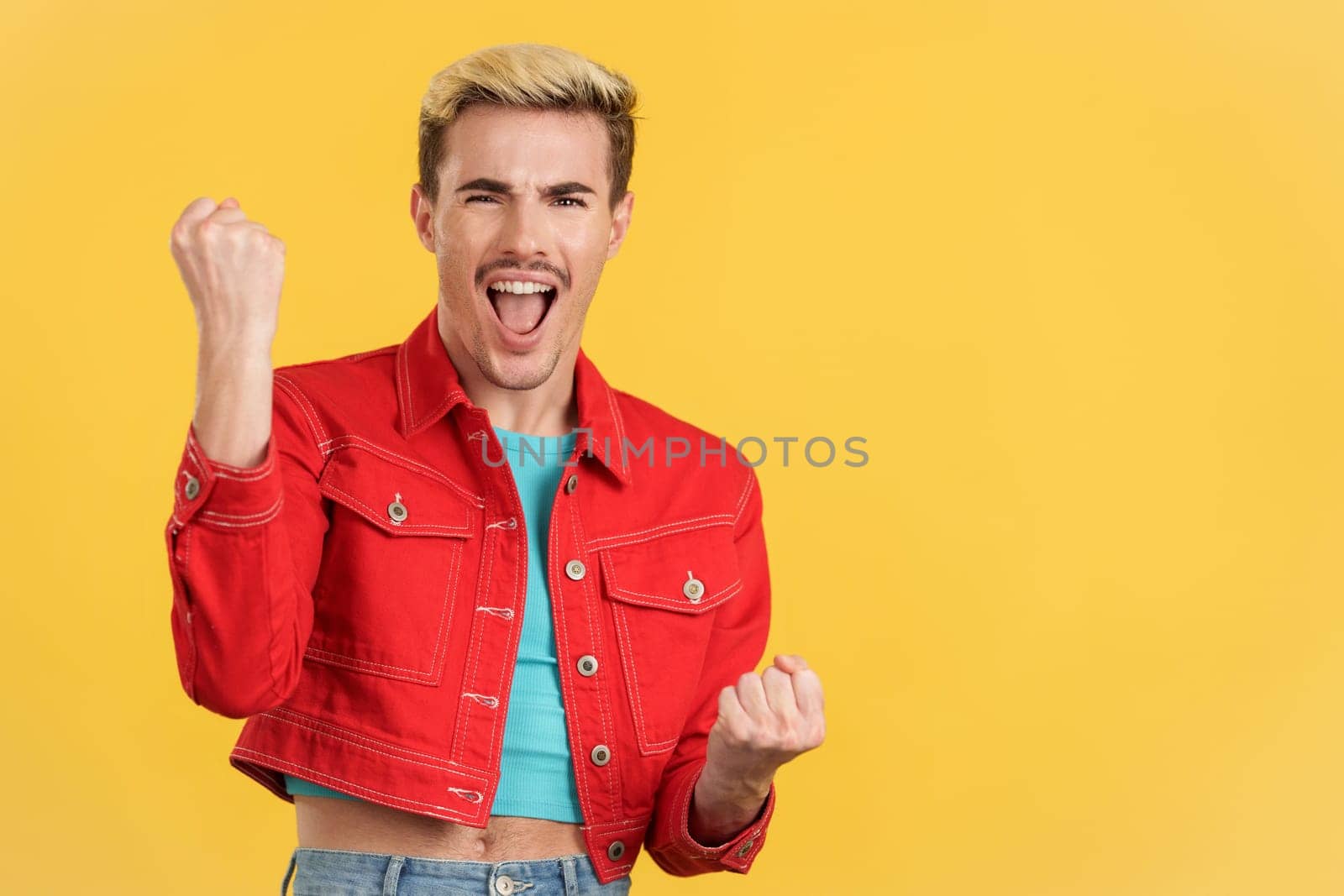 Happy gay man celebrating while raising fists in studio with yellow background