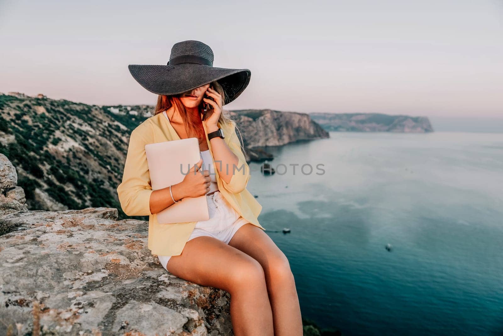 Successful business woman in yellow hat working on laptop by the sea. Pretty lady typing on computer at summer day outdoors. Freelance, travel and holidays concept.