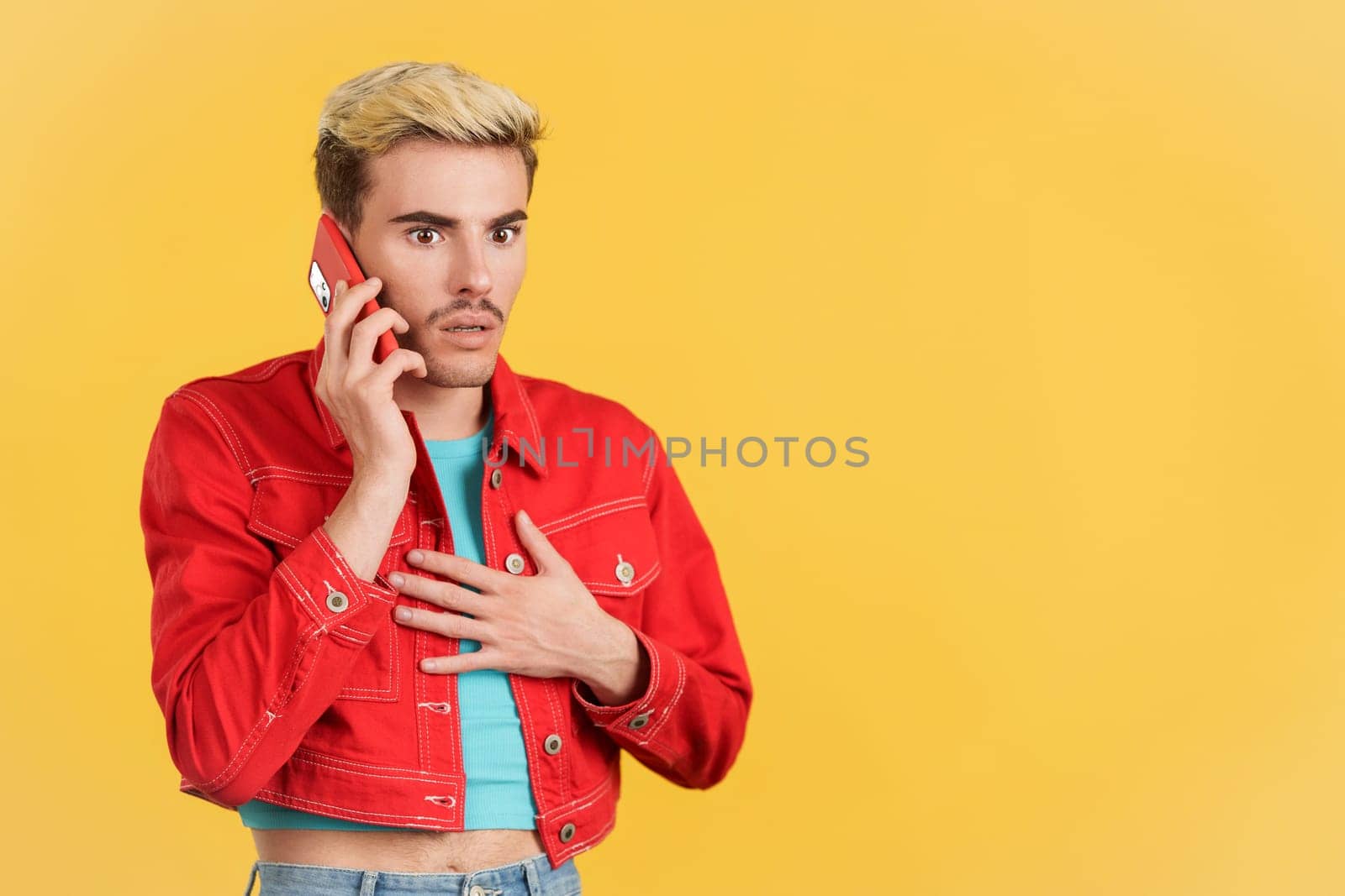 Astonished gay man talking to the mobile in studio with yellow background