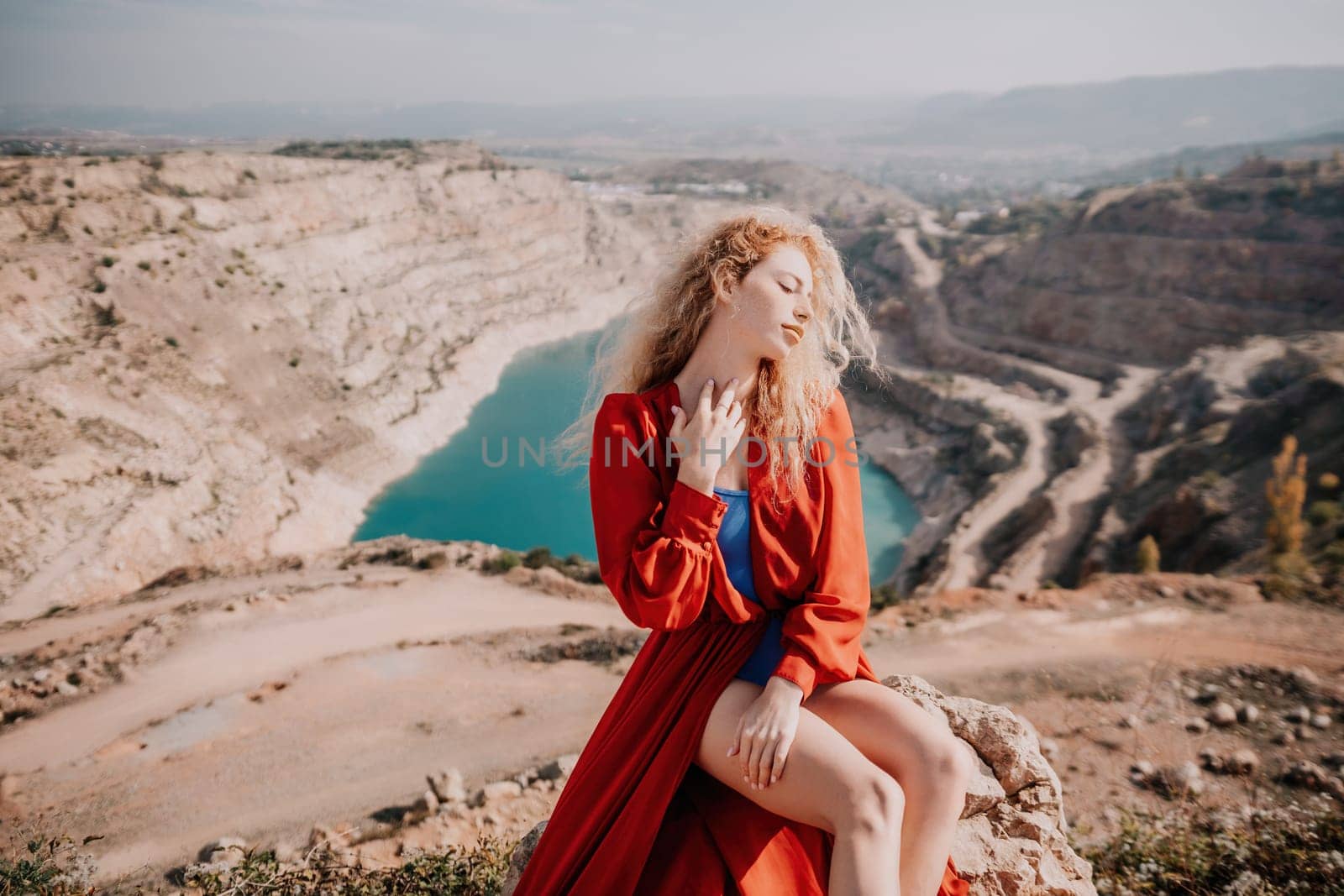 Close up portrait of curly redhead young caucasian woman with freckles looking at camera and smiling. Cute woman portrait in a pink long dress posing on a volcanic rock high above the sea at sunset by panophotograph