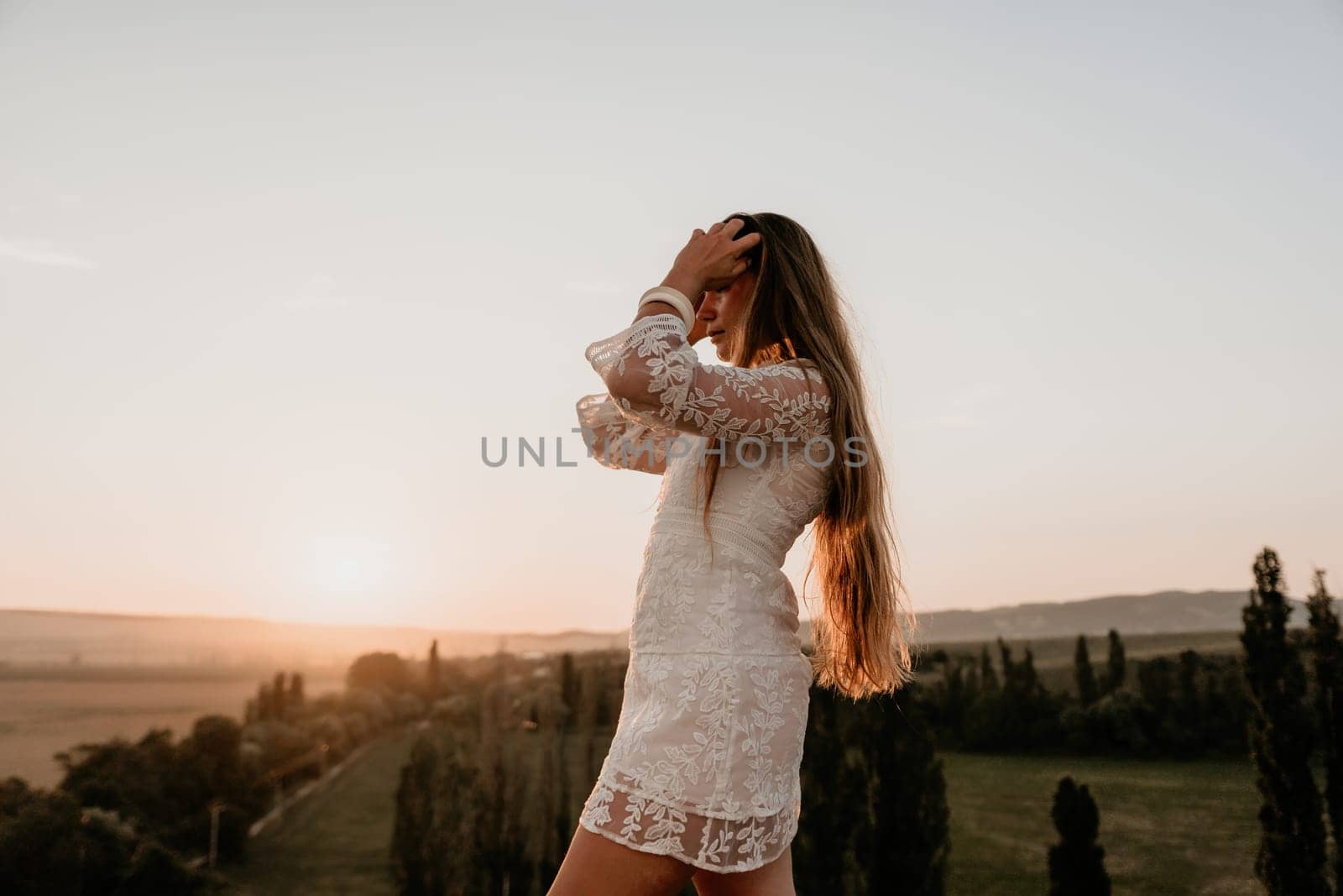 Happy woman in white boho dress on sunset in mountains. Romantic woman with long hair standing with her back on the sunset in nature in summer with open hands. Silhouette. Nature. Sunset. by panophotograph