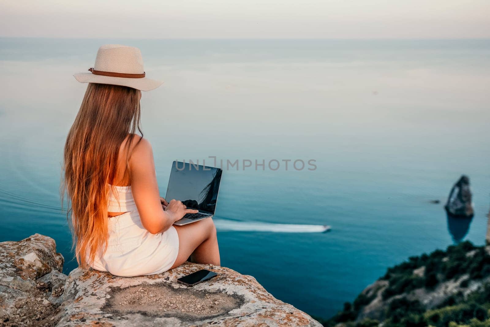 Woman laptop sea. Working remotely on seashore. Happy successful woman female freelancer in straw hat working on laptop by the sea at sunset. Freelance, remote work on vacation by panophotograph