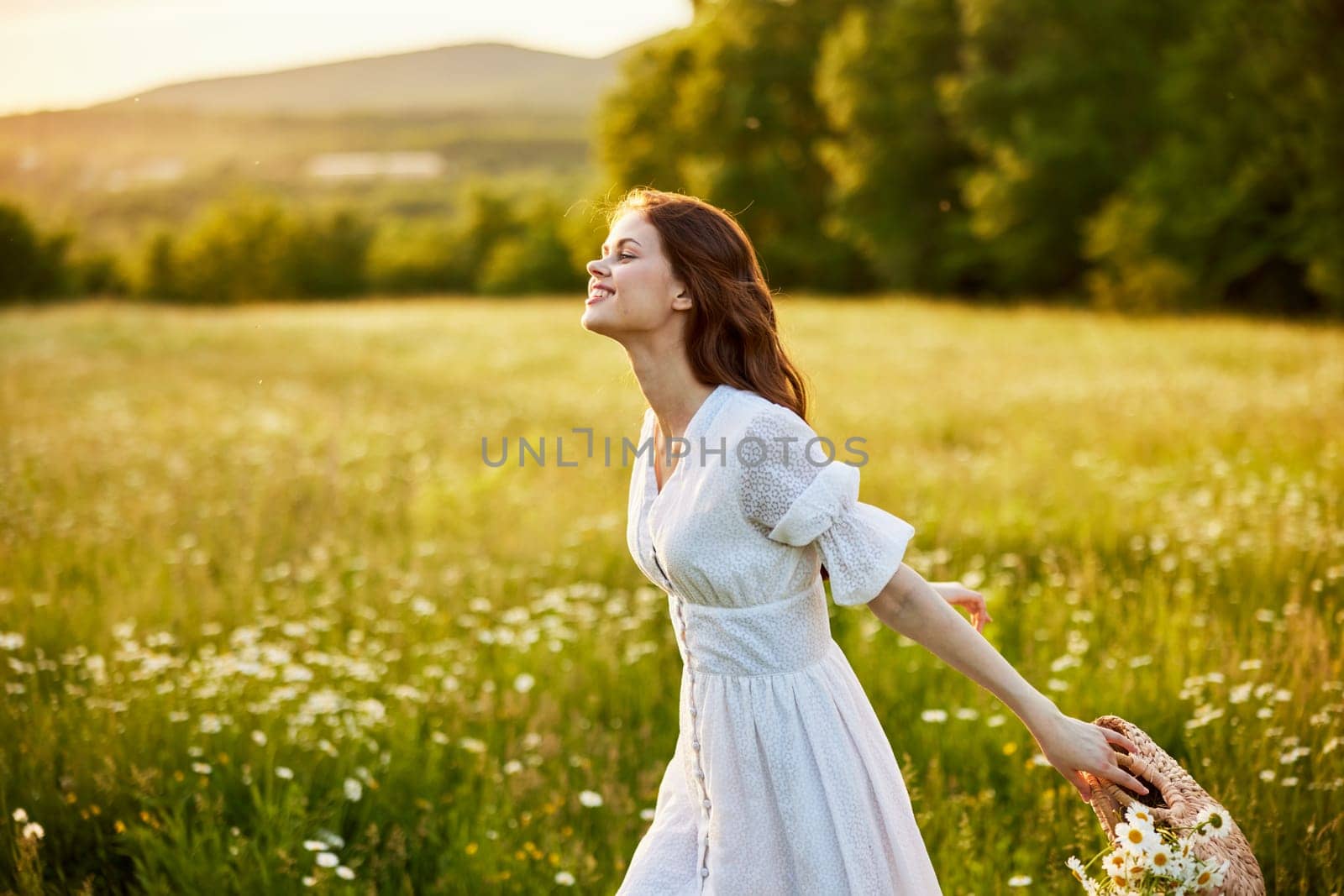 happy woman in a light dress and a wicker basket full of daisies enjoys nature walking in the field by Vichizh