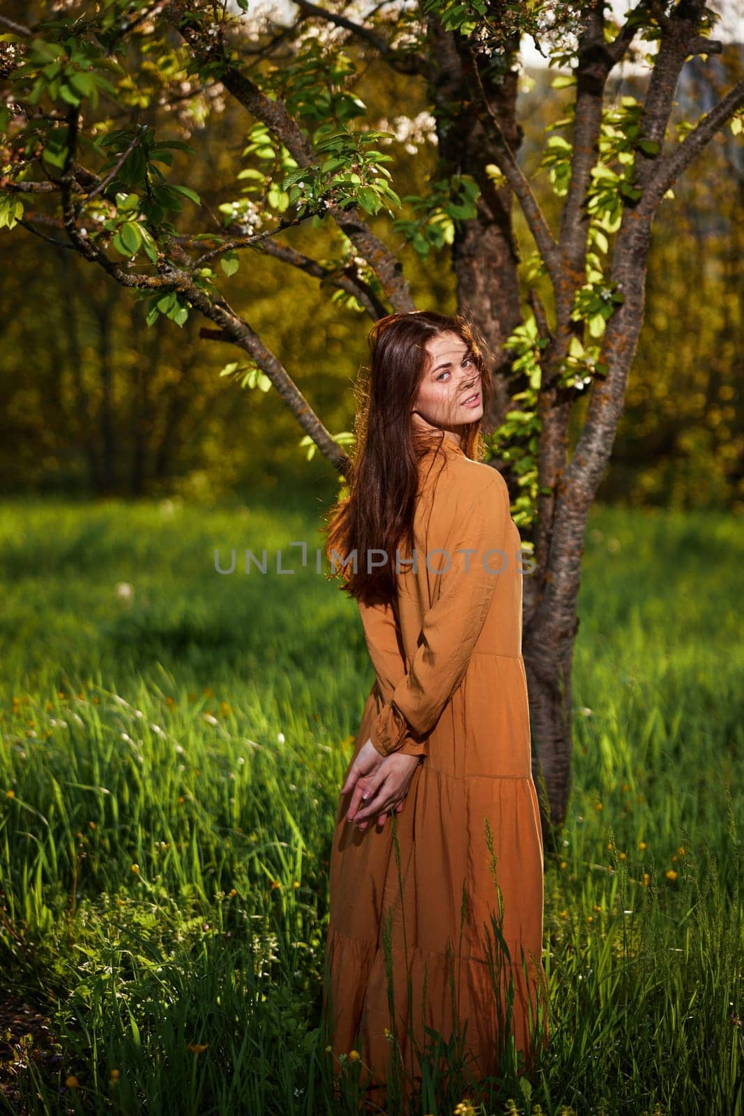 attractive woman with long red hair stands in the countryside near a flowering tree in a long orange dress and looks at the camera over her shoulder, with her hands folded on her back. by Vichizh