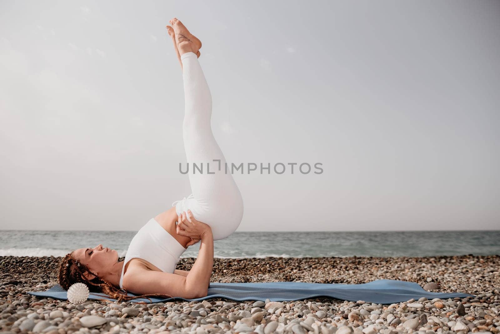 Woman yoga sea. Well looking middle aged woman with braids dreadlocks in white leggings and tops doing stretching pilates on yoga mat near sea. Female fitness yoga routine concept. Healthy lifestyle