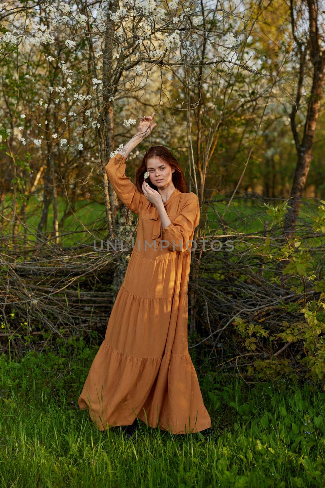 an elegant, sophisticated woman poses standing near a wicker fence at the dacha in a long orange dress by Vichizh