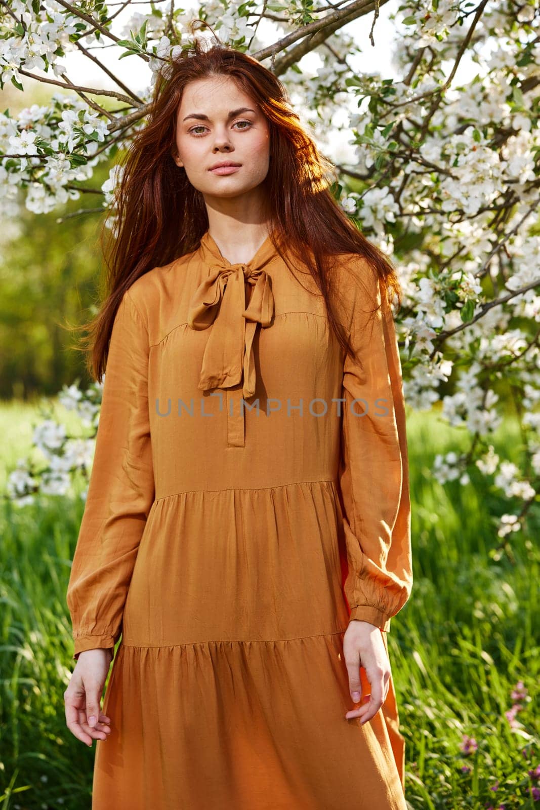 a bright vertical photo of an attractive woman in a long, stylish orange dress standing next to a flowering tree in sunny, warm weather, looking pleasantly into the camera, illuminated from the side by Vichizh