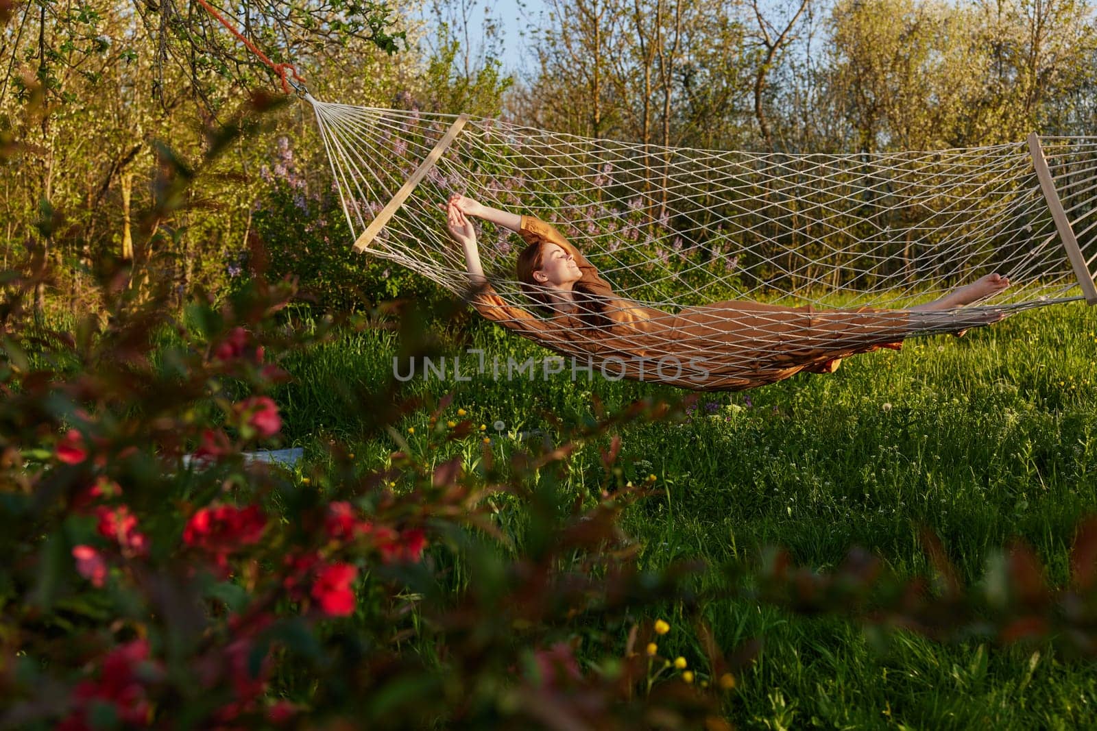 a happy woman in a long orange dress is relaxing in nature lying in a mesh hammock enjoying the peace and tranquility in the country surrounded by green foliage by Vichizh
