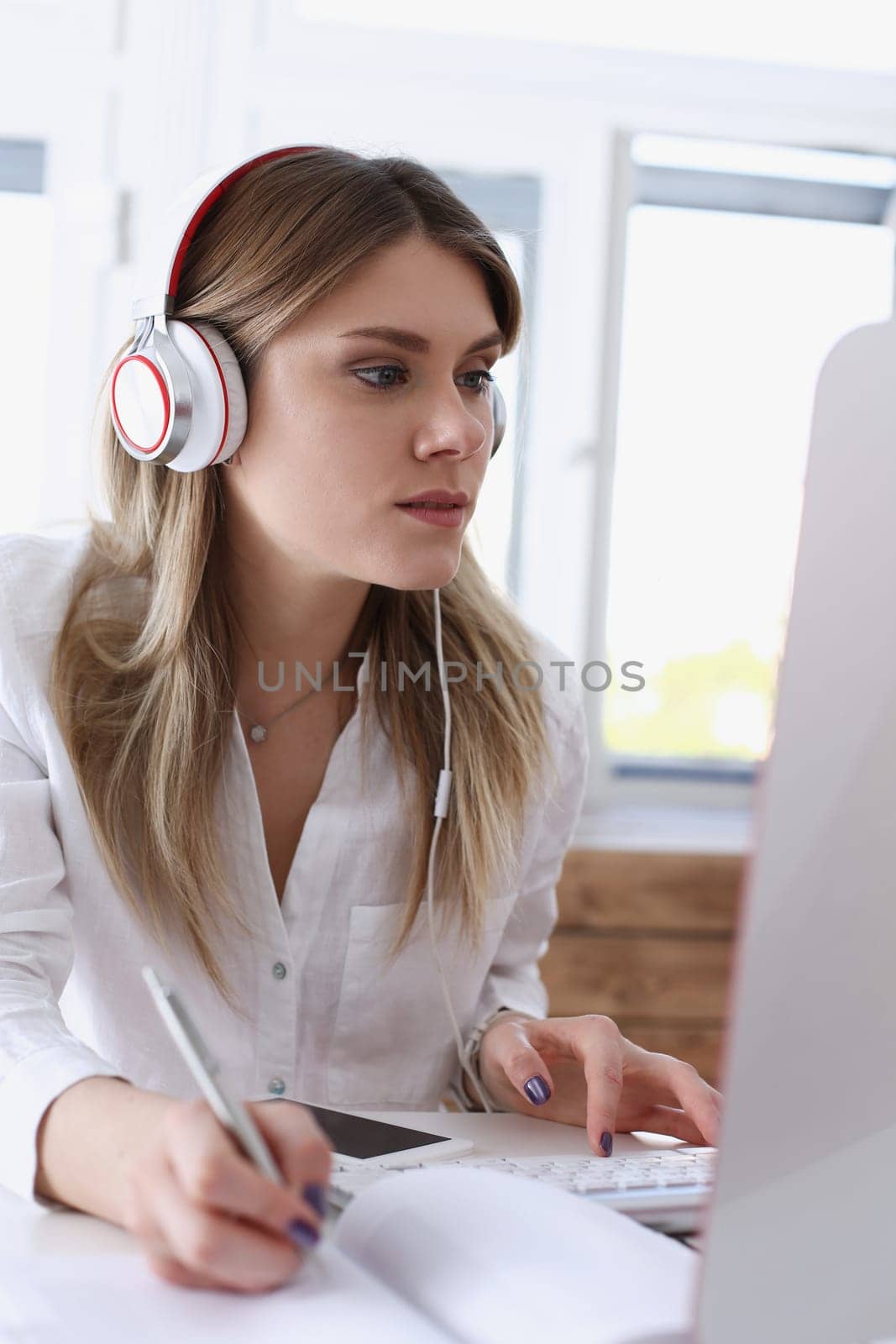 Beautiful woman wearing headphones making notes with silver pen while working on computer pc portrait. Management training course modern remote school self development and perfection concept