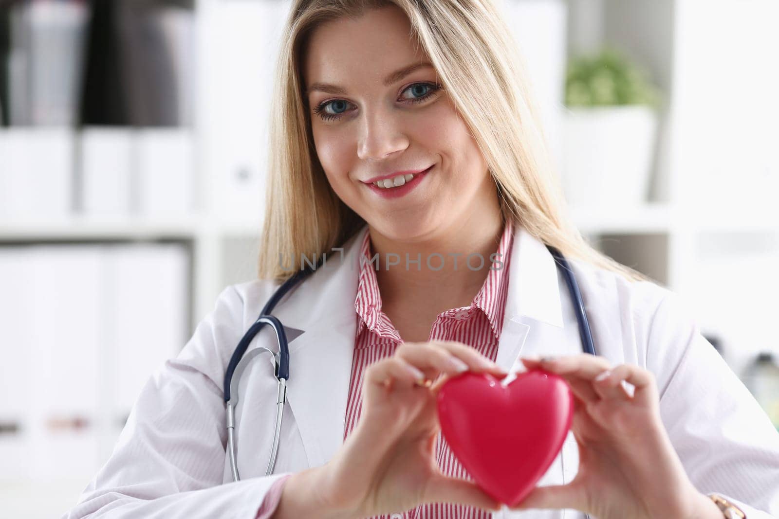 Beautiful smiling blond female doctor hold in arms red toy heart closeup. Cardio therapeutist student education CPR 911 life save physician make cardiac physical pulse rate measure arrhythmia