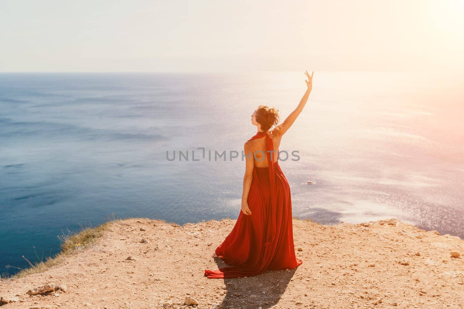 Side view a Young beautiful sensual woman in a red long dress posing on a rock high above the sea during sunrise. Girl on the nature on blue sky background. Fashion photo.