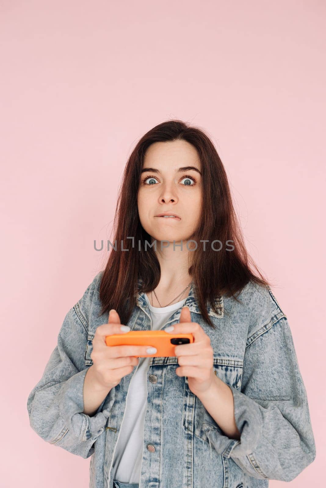 Portrait of a young woman in casual denim jacket with smartphone playing game, isolated on pink background conveying hesitancy and insecurity.