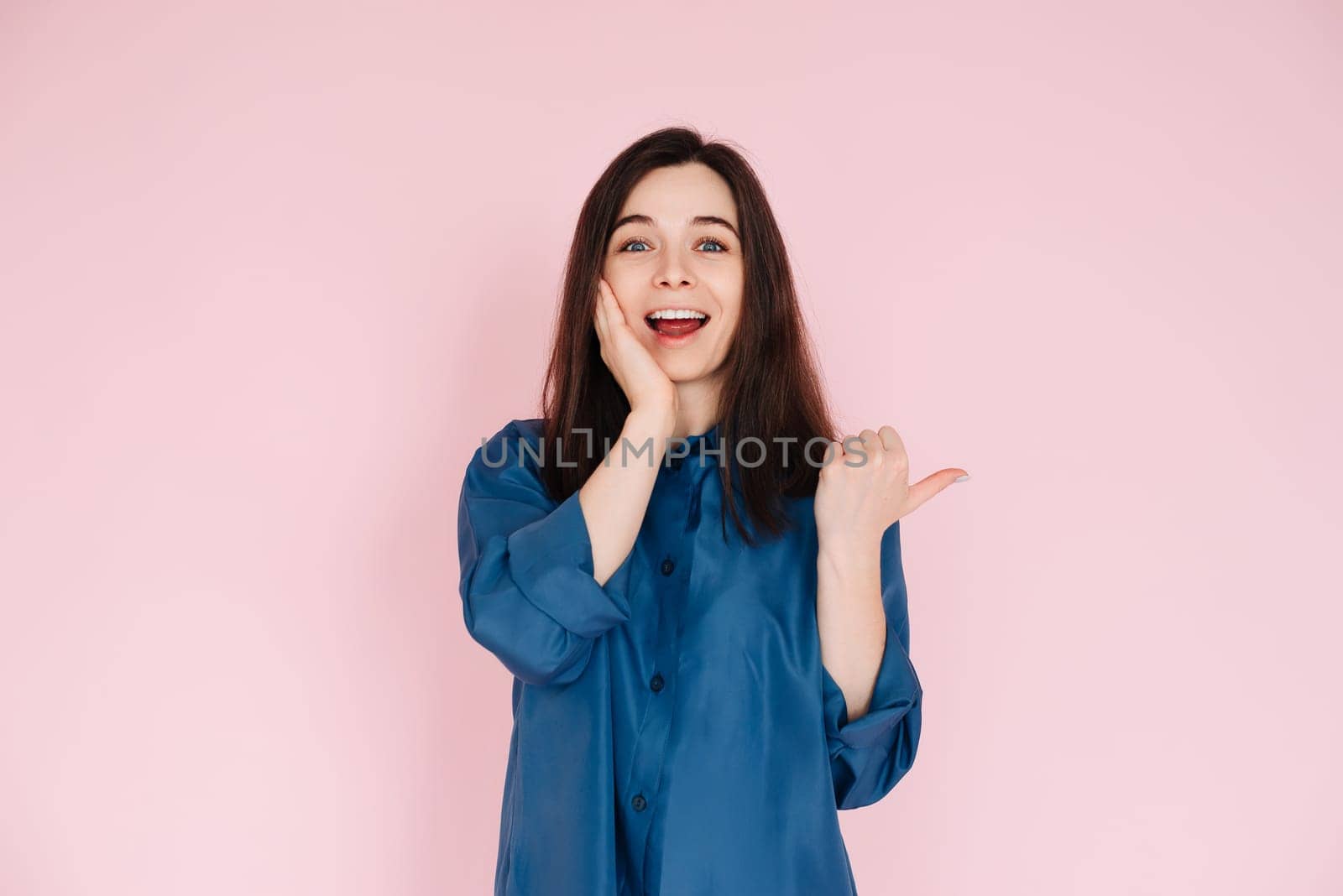 Young woman expressing amazement and joy, touching cheek and pointing finger at empty space, with a big toothy smile on pink isolated background.