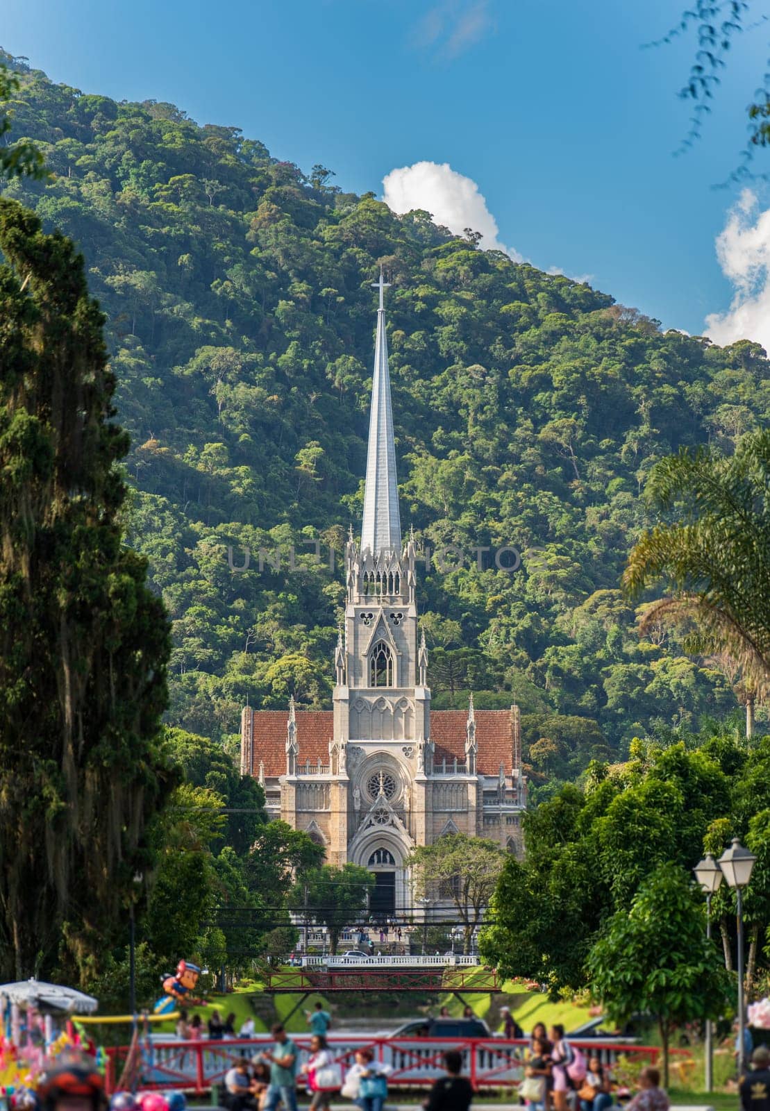 Impressive view of the towering Petropolis Cathedral in the distance, with a dense jungle mountain range as a stunning backdrop. People are walking around, but their figures are blurred, giving an ethereal quality to the scene.