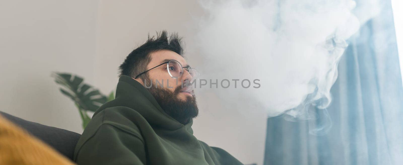 Bearded man is smoking hookah at home and blowing cloud of smoke - chill time and resting