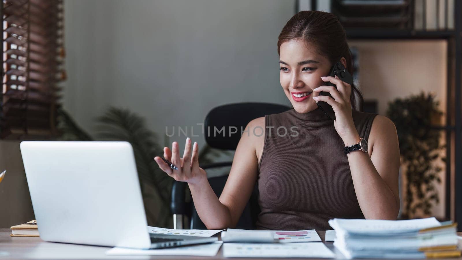 Smiling Asian Accountant talking phone with client and makes financial report and studies annual figures, analyzes profits.