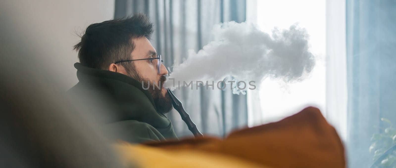 Bearded millennial or gen z man smoking hookah while relaxing on sofa at home - chill time and resting