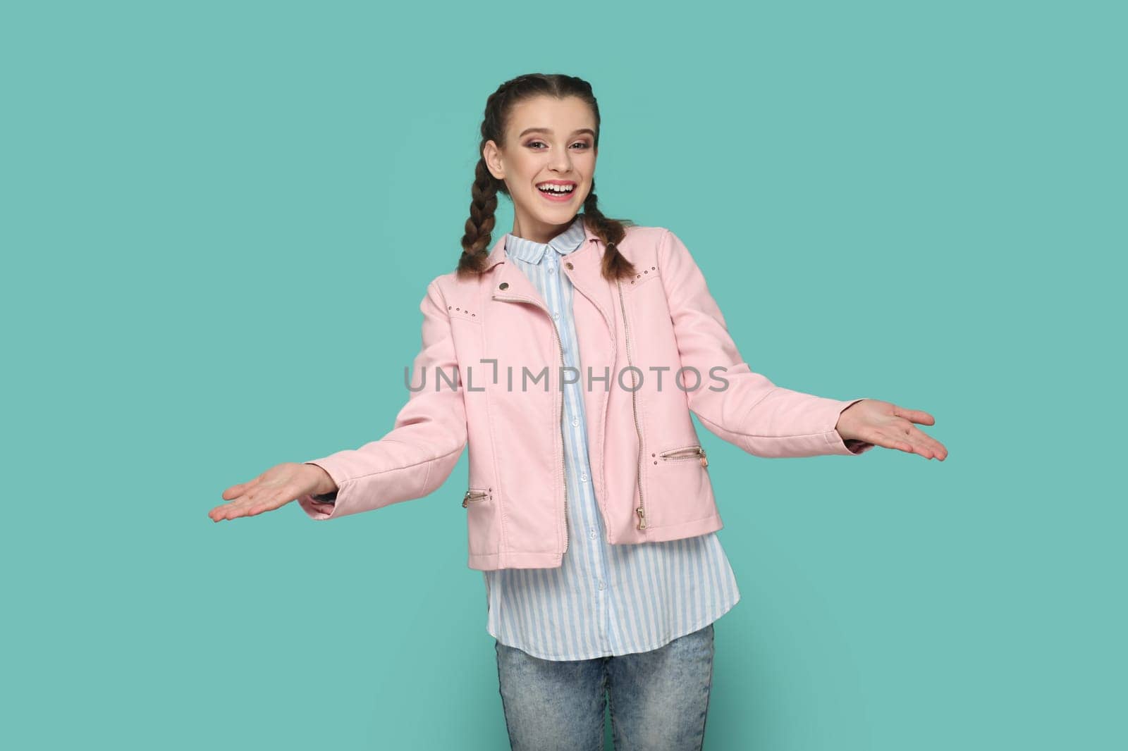 Portrait of friendly teenager girl with braids wearing pink jacket standing with open hands, sharing with somebody for free. Indoor studio shot isolated on green background.