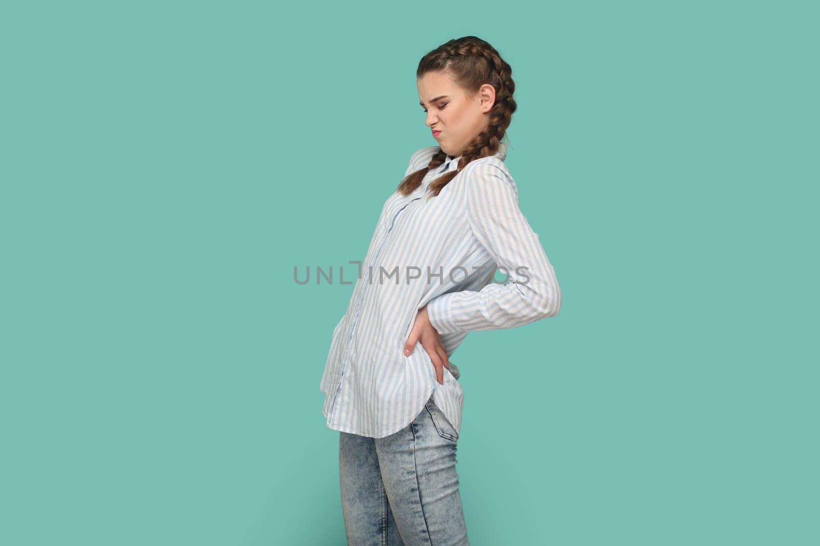 Portrait of dissatisfied teenager girl with braids wearing striped shirt smirks face, keeps hand on back, suffers from terrible pain. Indoor studio shot isolated on green background.