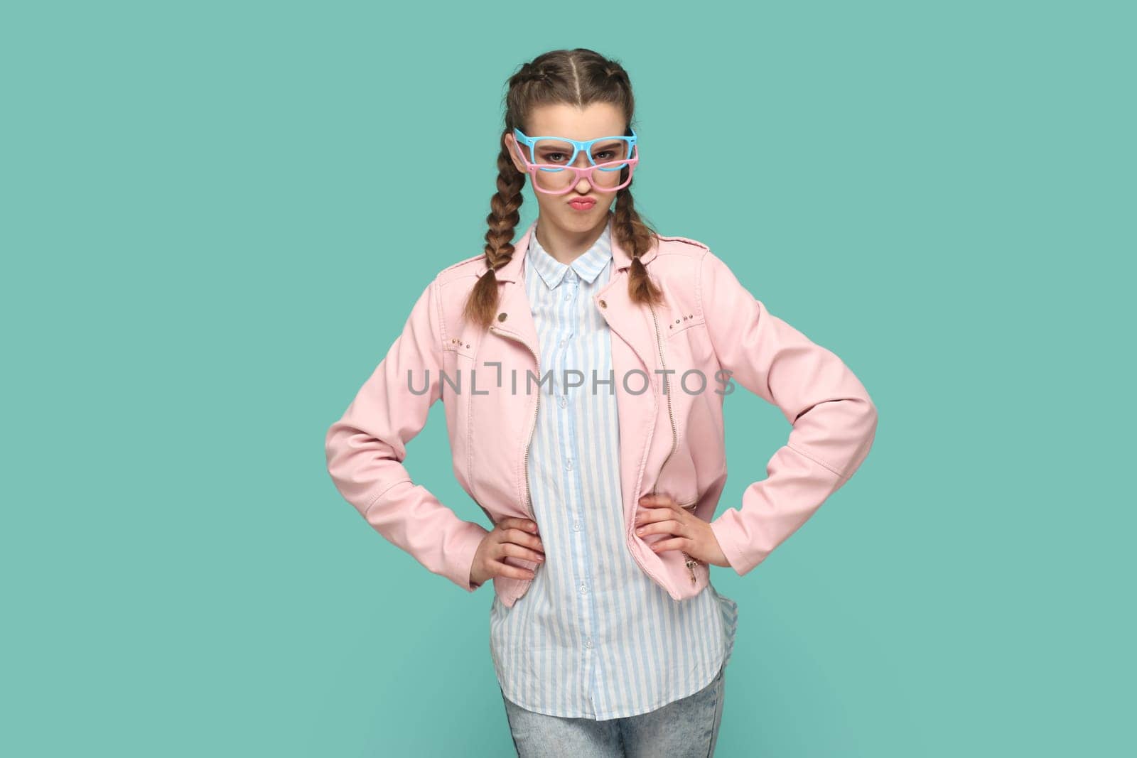 Portrait of confident funny teenager girl with braids wearing pink jacket standing with hands on hips, wearing two optical glasses, choosing best color. Indoor studio shot isolated on green background