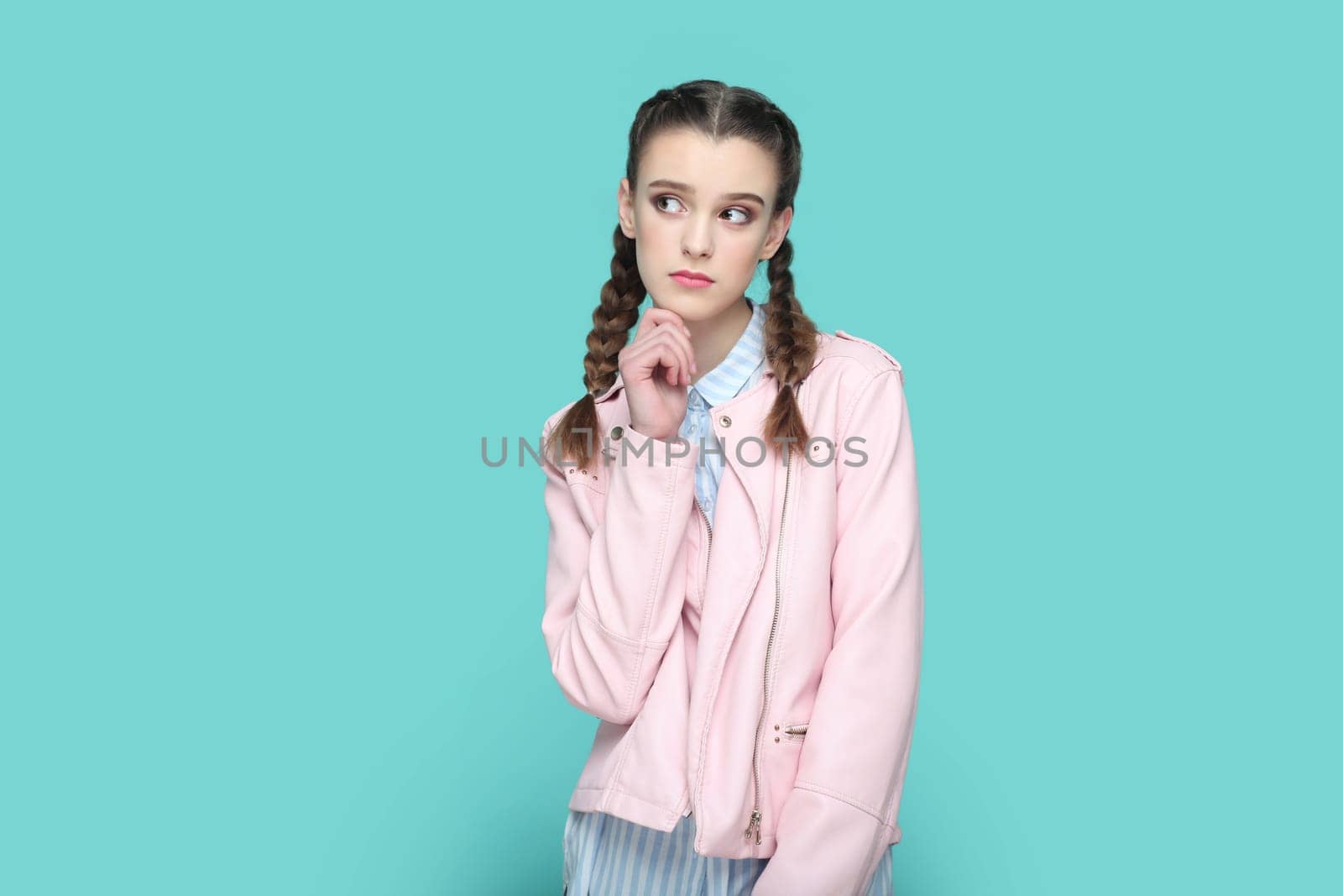 Portrait of pensive thoughtful teenager girl with braids wearing pink jacket standing holding her chin, looking away, planning her future. Indoor studio shot isolated on green background.