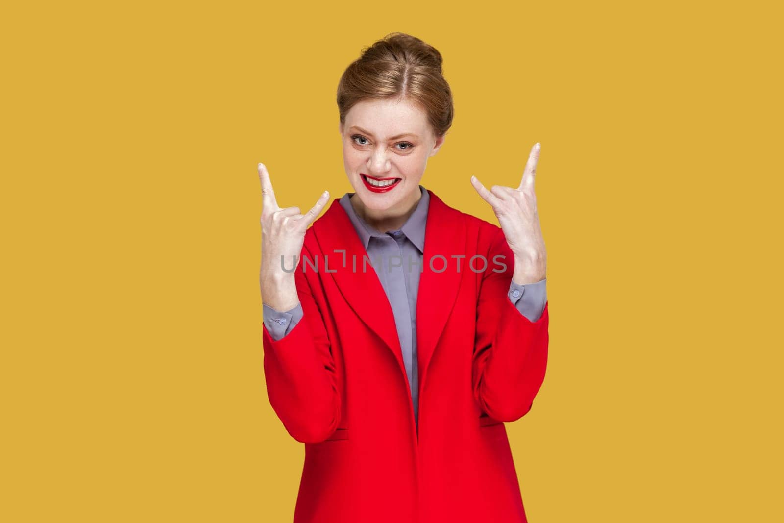 Portrait of excited crazy woman with red lips standing showing rock and roll gesture, having fun on festival, wearing red jacket. Indoor studio shot isolated on yellow background.