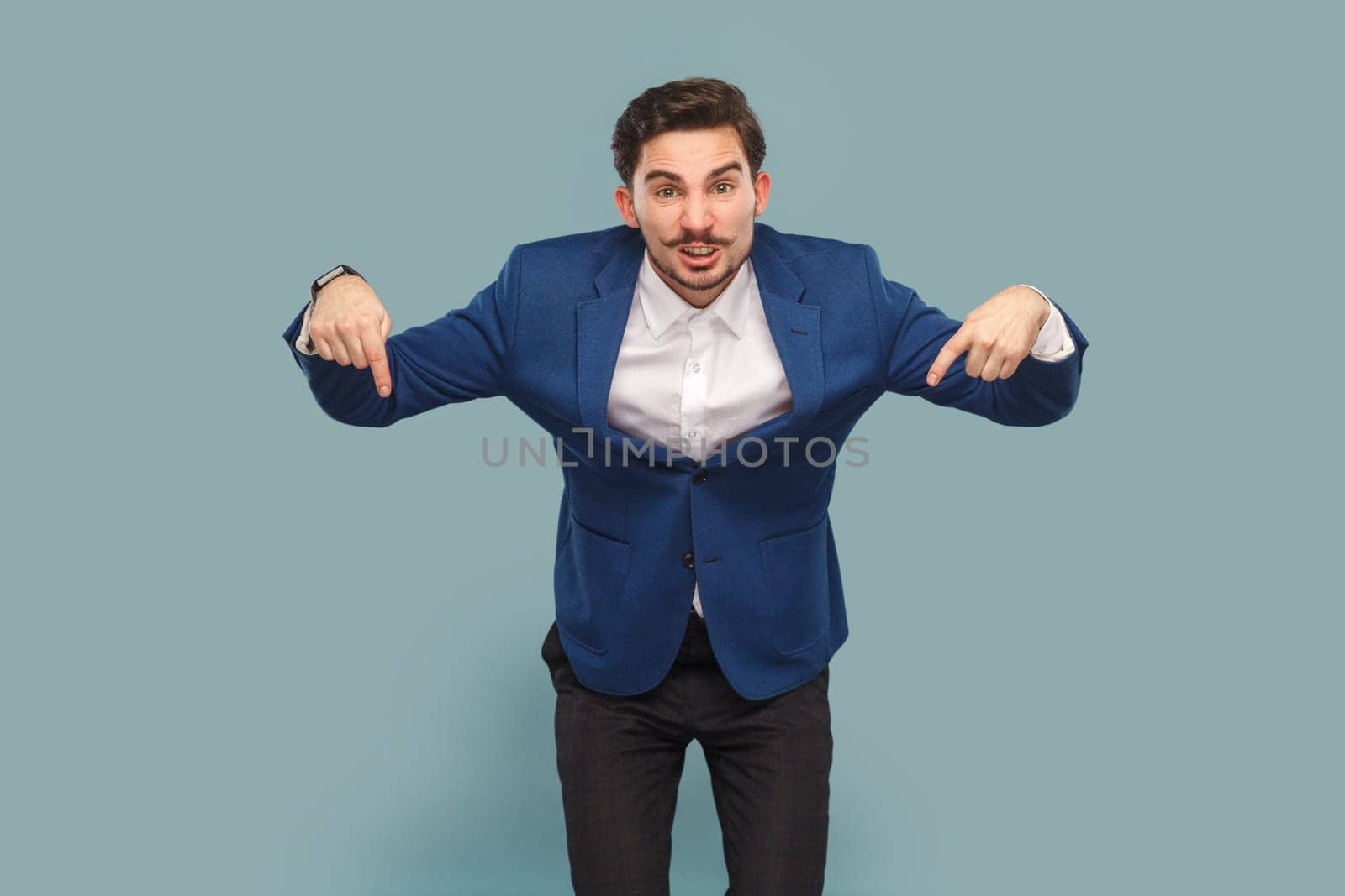 Angry man with mustache standing pointing down, giving command, looking at camera with serious expression, wearing white shirt and jacket. Indoor studio shot isolated on light blue background.