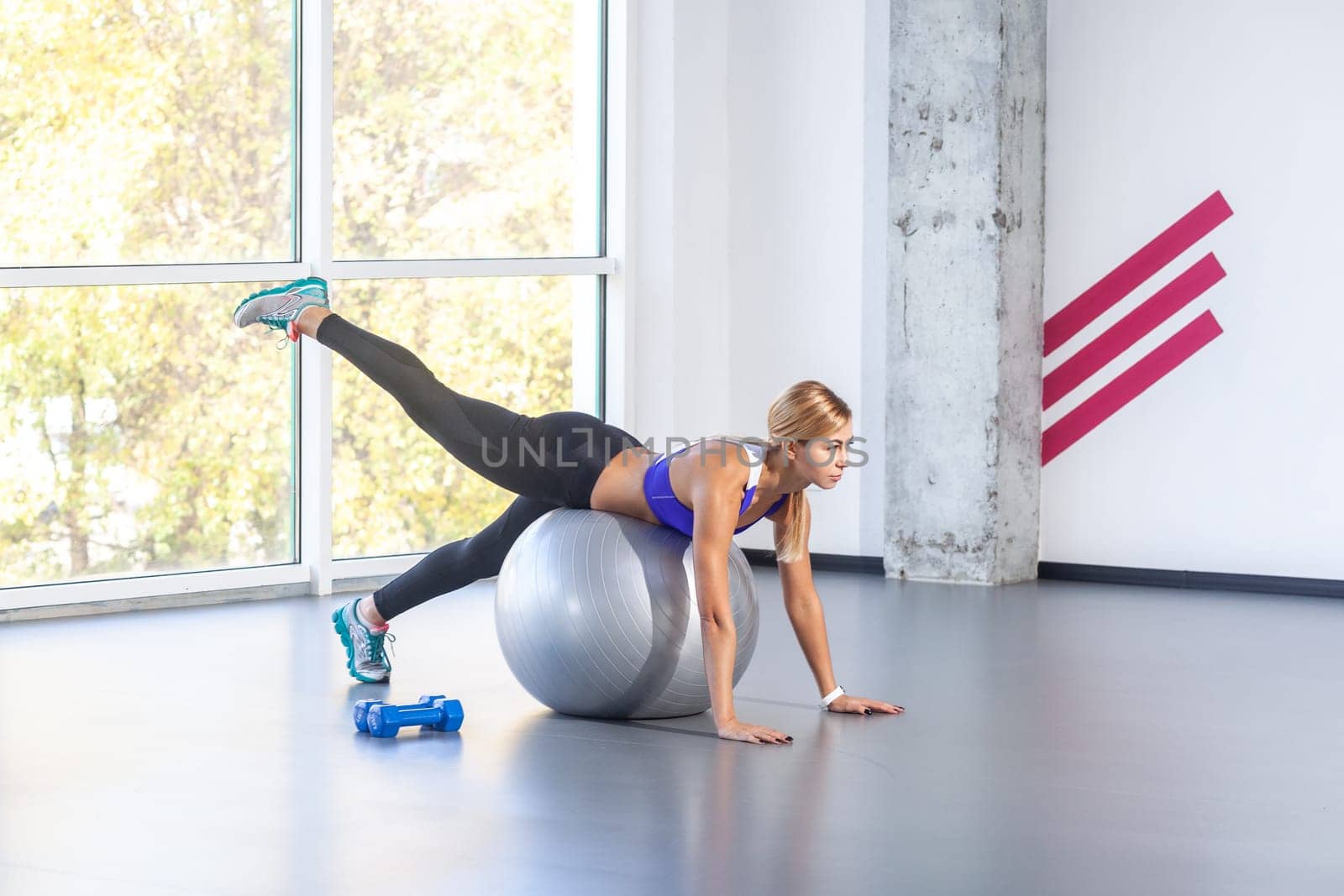 Side view portrait of slim attractive sporty woman lying on a fitness ball with her palms on floor, raised her leg up, wearing sports top and tights. Indoor shot with window on background.