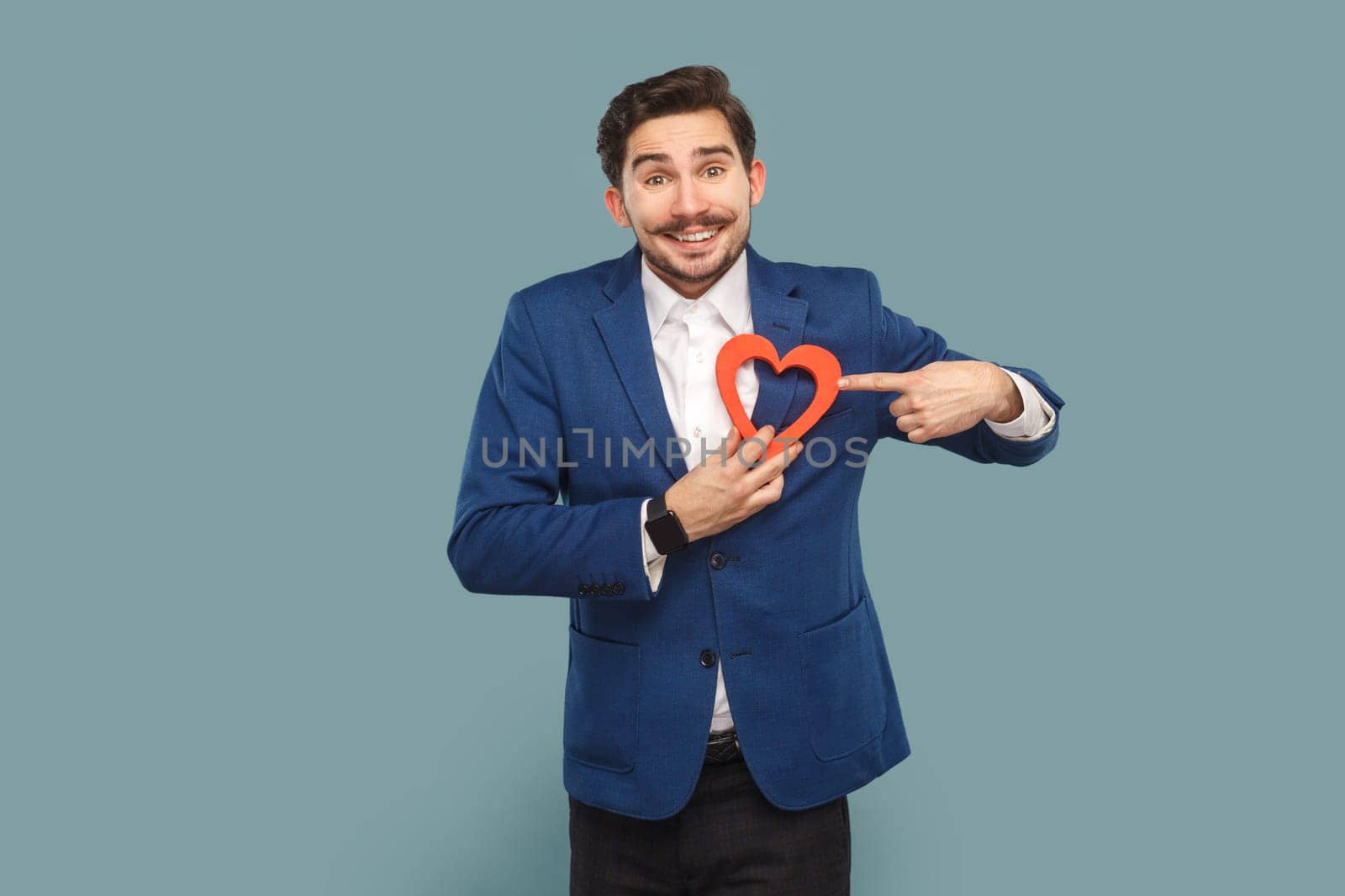 Portrait of funny positive optimistic man with mustache standing pointing at heart figure, looking at camera, wearing white shirt and jacket. Indoor studio shot isolated on light blue background.