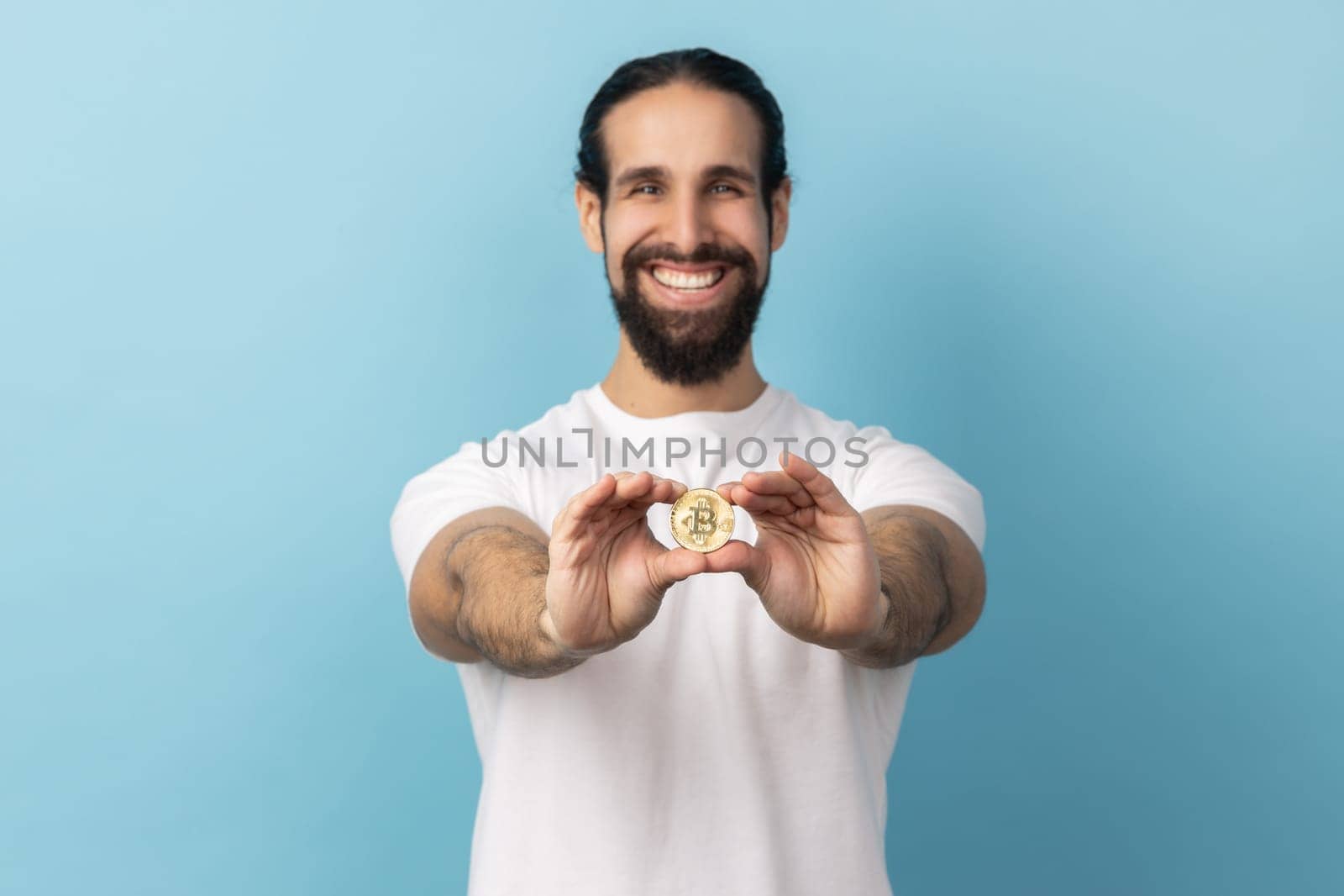 Portrait of satisfied man with beard wearing white T-shirt standing holding out bitcoin, paying attention at new digital cryptocurrency. Indoor studio shot isolated on blue background.