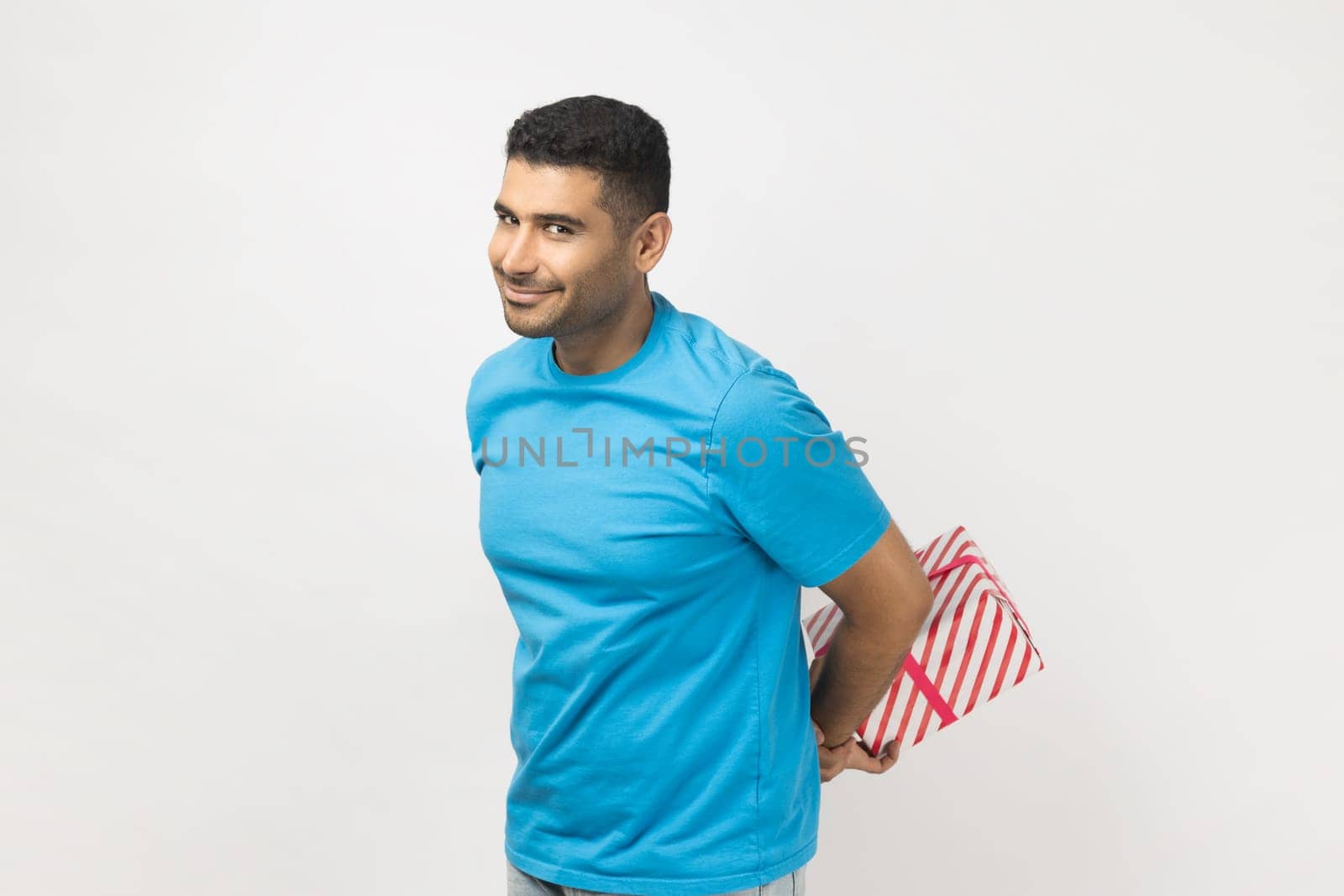 Portrait of positive cheerful unshaven man wearing blue T- shirt standing with present box behind back, gift for holiday, congratulating. Indoor studio shot isolated on gray background.