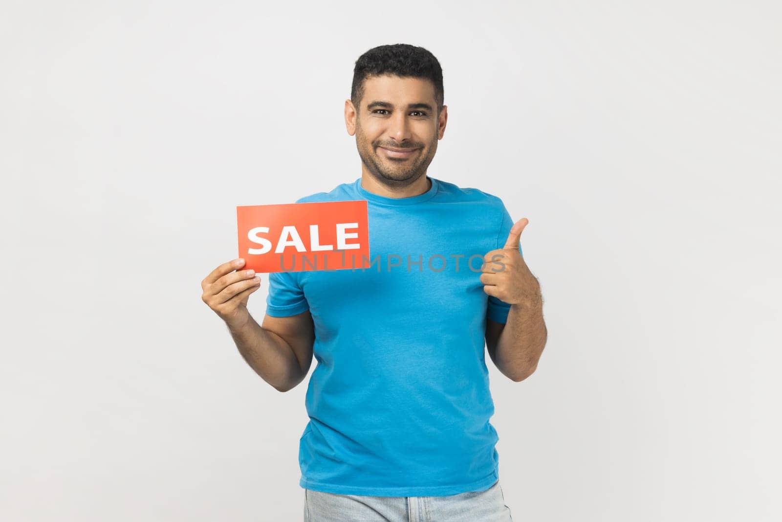 Portrait of cheerful joyful man wearing blue T- shirt standing holding sale card in hands looking at camera with toothy smile, showing thumb up. Indoor studio shot isolated on gray background.