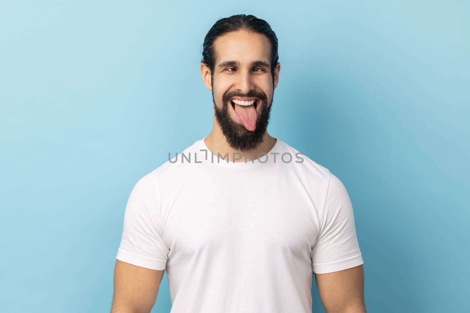 Portrait of comic positive man with beard wearing white T-shirt looking cross-eyed, having fun with silly face expression, showing tongue out. Indoor studio shot isolated on blue background.