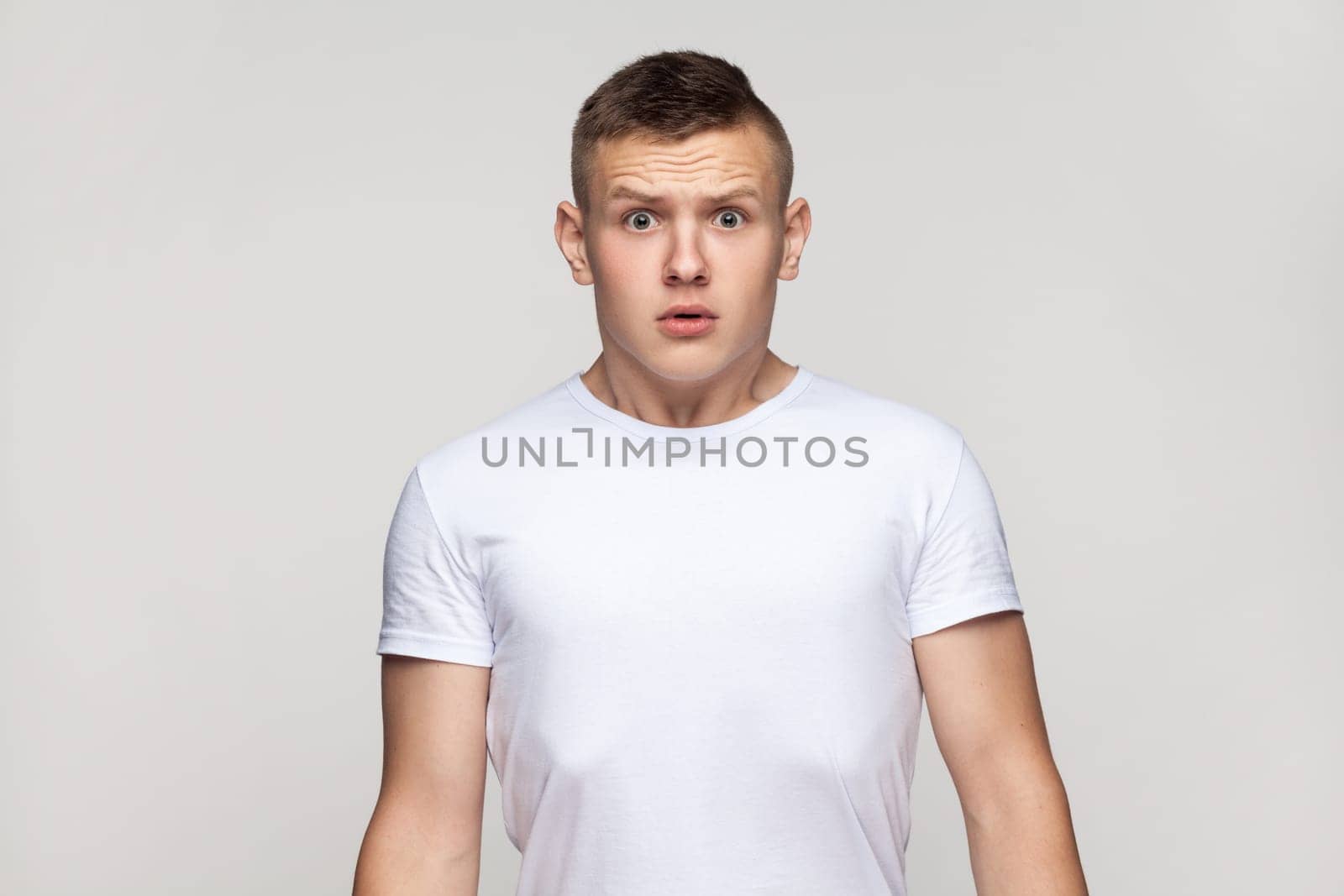 Portrait of astonished surprised attractive teenager boy wearing T-shirt standing looking at camera with big eyes, being frighten and scared. Indoor studio shot isolated on gray background.