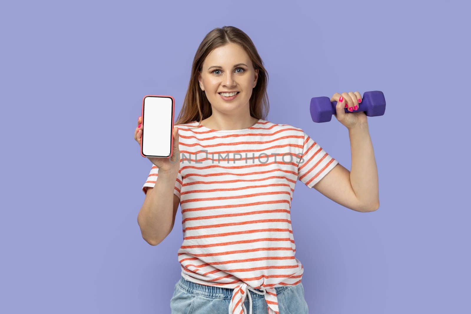 Portrait of delighted good looking woman wearing striped T-shirt holding dumbbell in hand, showing smart phone with empty display for advertisement. Indoor studio shot isolated on purple background.