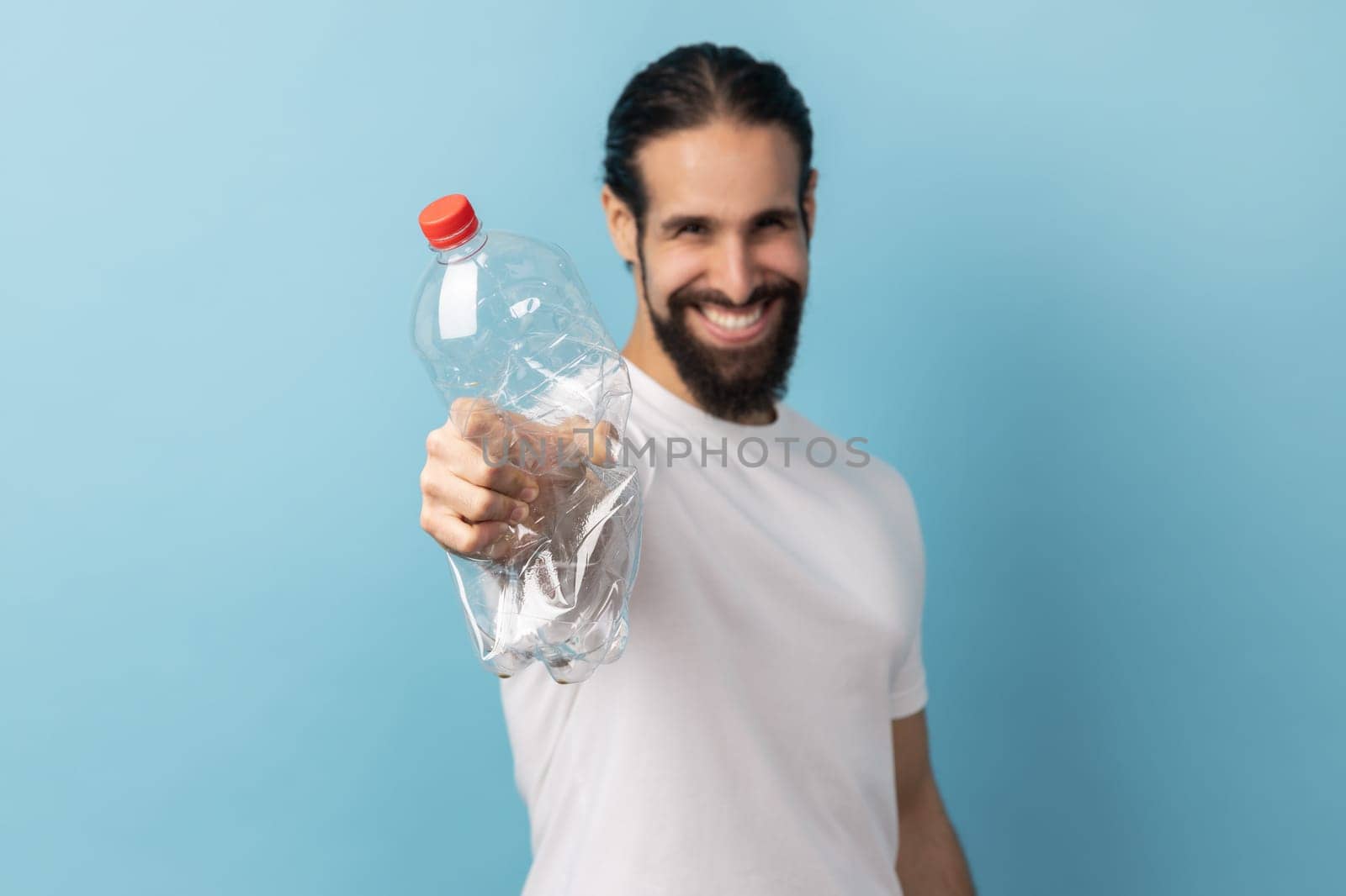 Portrait of smiling man with beard wearing white T-shirt holding out plastic bottle, worrying about plastic recycling, environmental pollution. Indoor studio shot isolated on blue background.