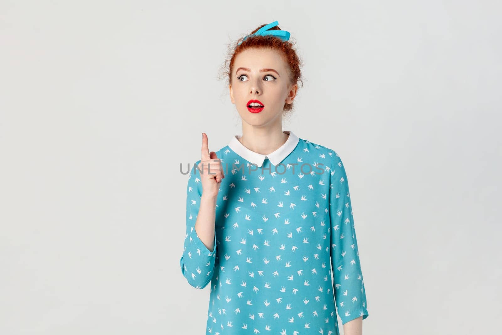 Portrait of excited clever smart red haired woman with bun hairstyle, standing raised index finger up, having idea, looking away, wearing blue dress. Indoor studio shot isolated on gray background.