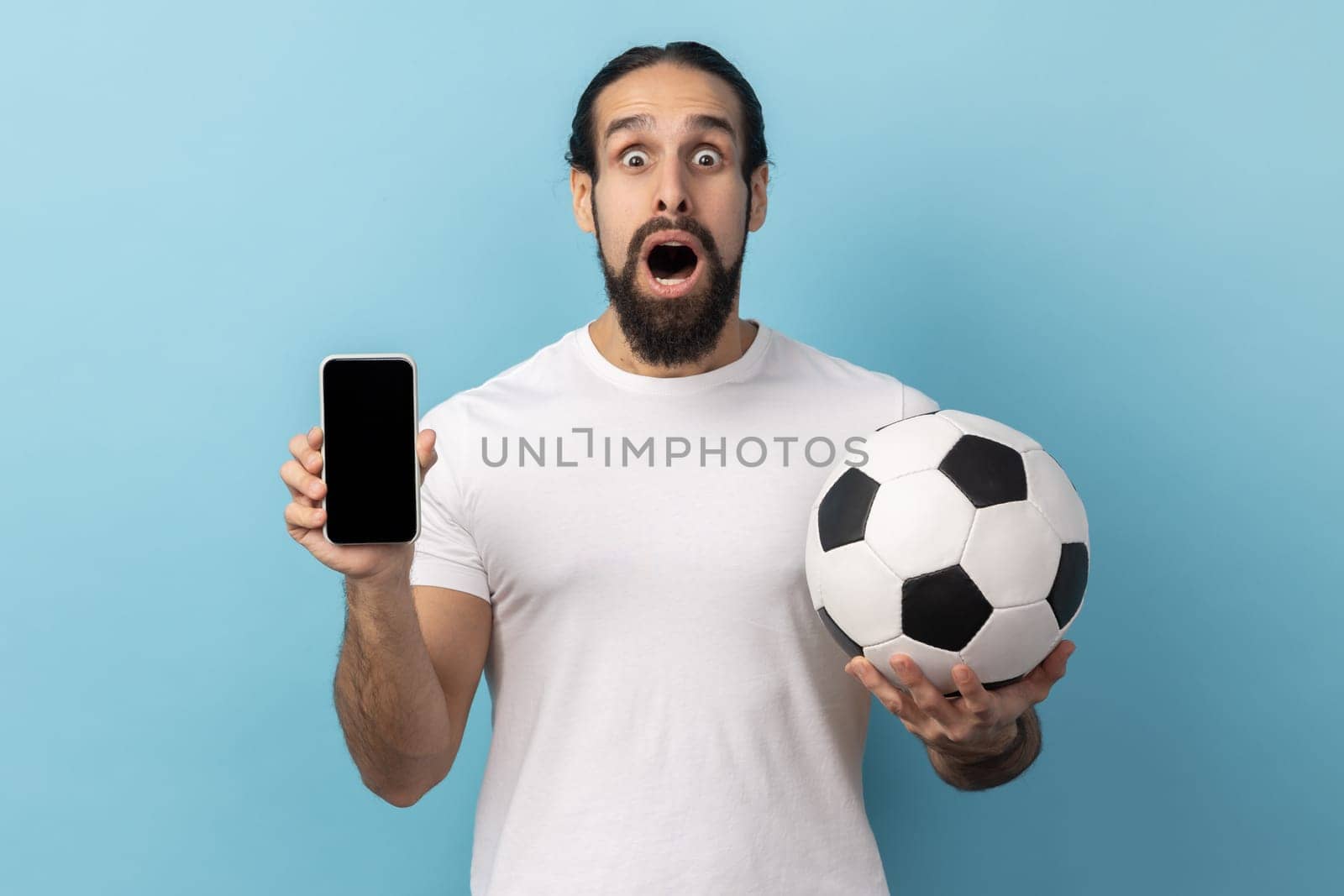 Portrait of amazed man with beard wearing white T-shirt holding soccer ball and smartphone empty black display, ticket booking for championship. Indoor studio shot isolated on blue background.