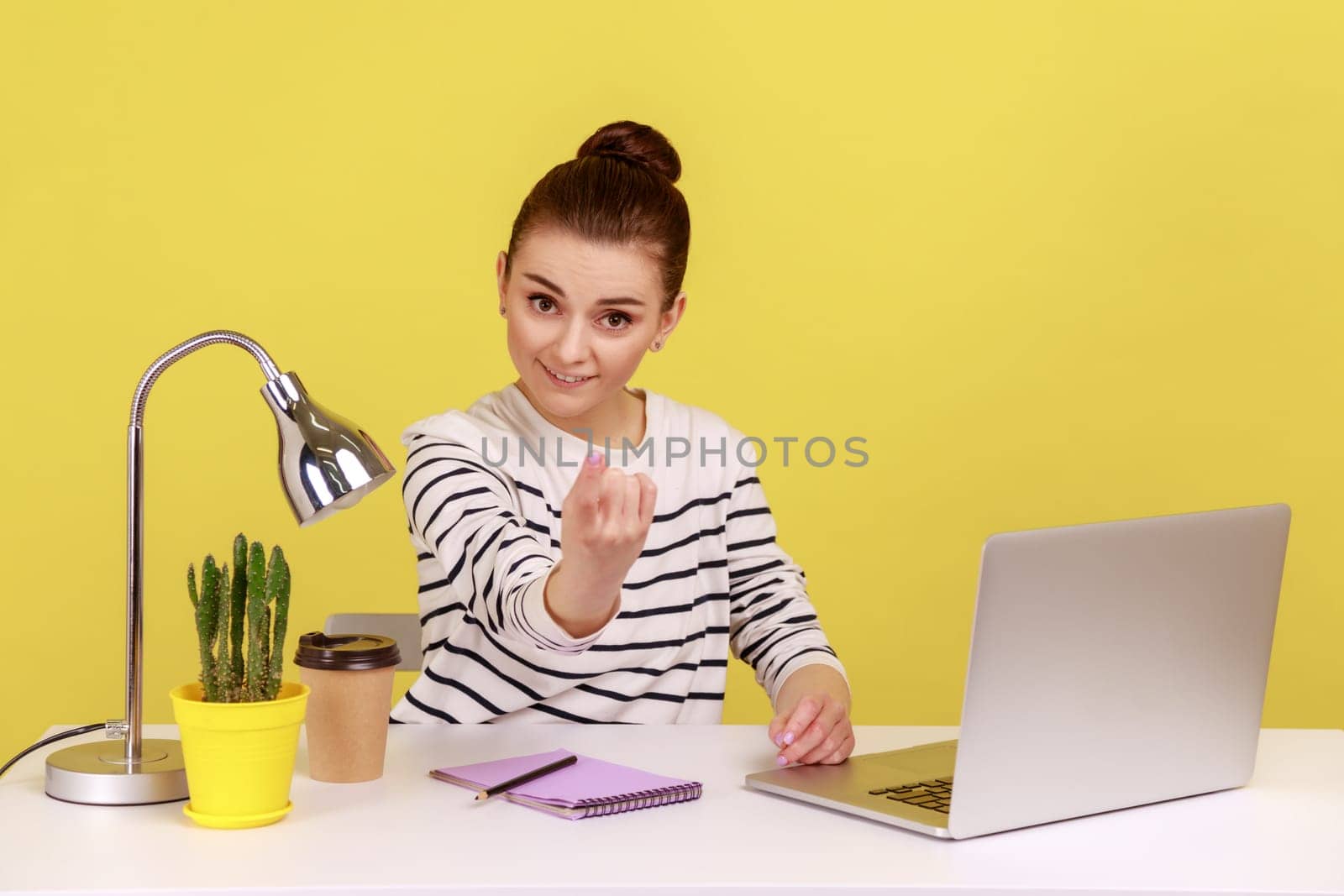 Come here. Woman manager sitting at workplace with laptop and making beckoning gesture, inviting to come hither, offering job application. Indoor studio studio shot isolated on yellow background.