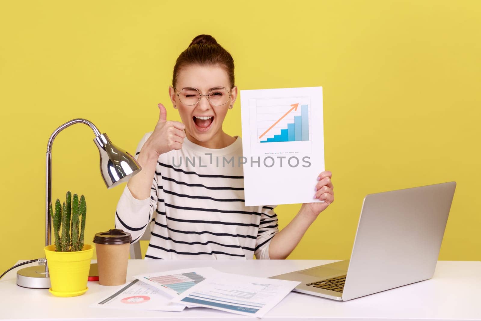 Delighted woman smiling at camera holding growth diagram, showing thumb up, satisfied with financial and economic growth of his business. Indoor studio studio shot isolated on yellow background.