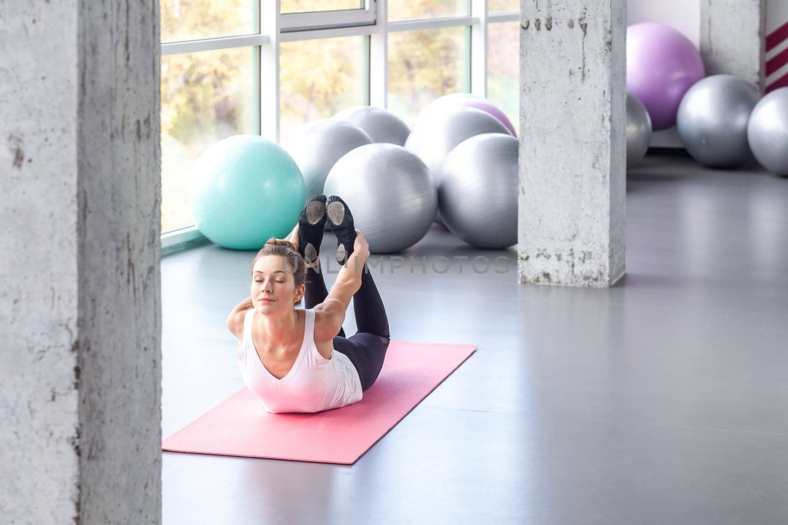 Portrait of sporty and flexible woman lying on bellies and holding legs in hands while practicing yoga on exercise mat, keeps eyes closed. Indoor shot with window on background.