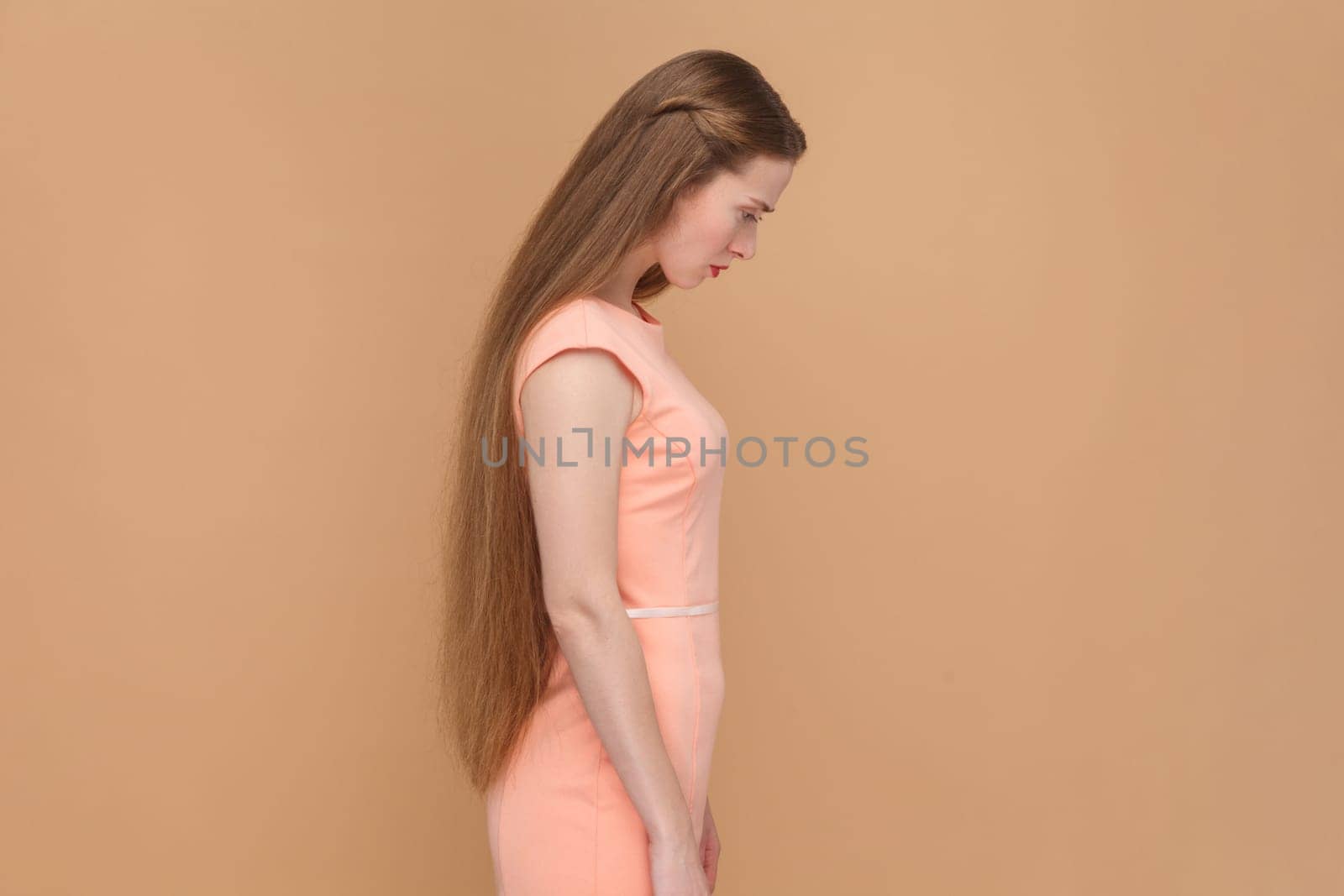 Side view portrait of sad upset woman with long hair hearing bad news, posing with head down, crying, expressing sorrow, wearing elegant dress. Indoor studio shot isolated on brown background.