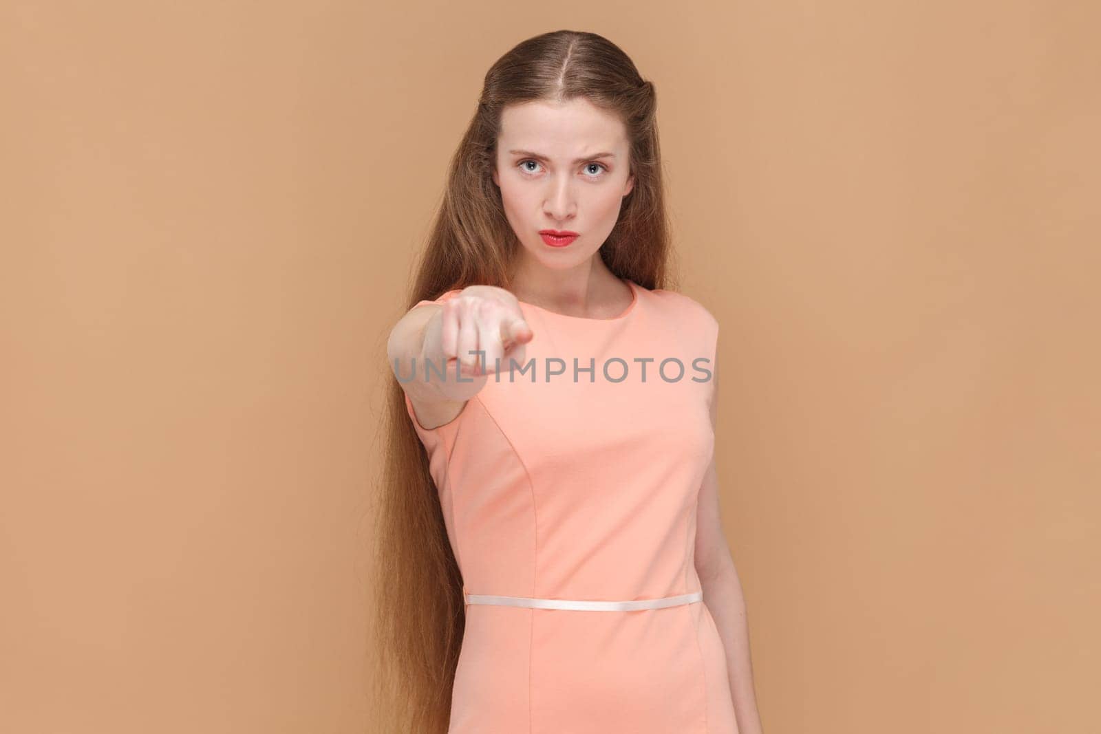 Portrait of serious strict woman with long hair standing pointing to camera, choosing you, select, making choice, wearing elegant dress. Indoor studio shot isolated on brown background.