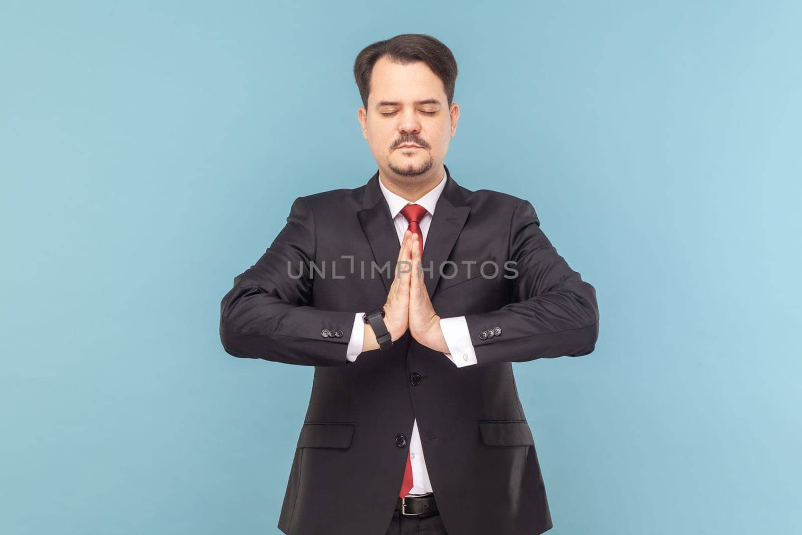 Portrait of calm man with mustache standing with closed practicing yoga, meditating while having break at work, wearing black suit with red tie. Indoor studio shot isolated on light blue background.