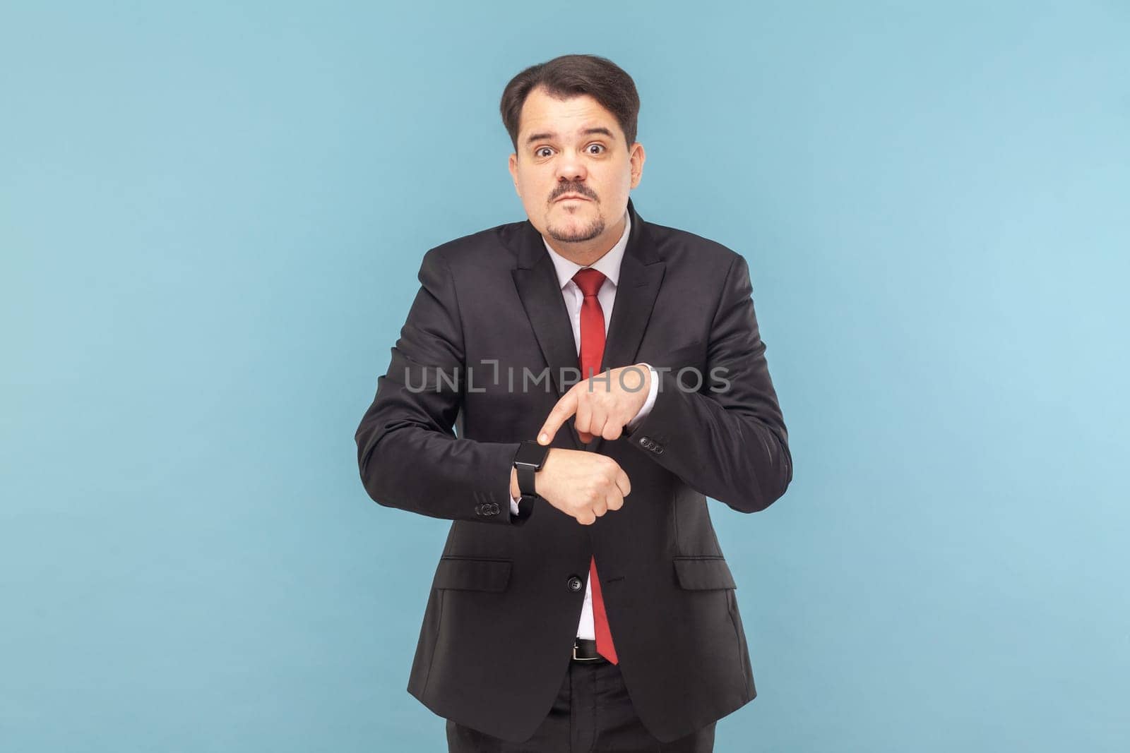 Portrait of serious man with mustache standing looking at camera, pointing at his smartwatch, deadline, wearing black suit with red tie. Indoor studio shot isolated on light blue background.