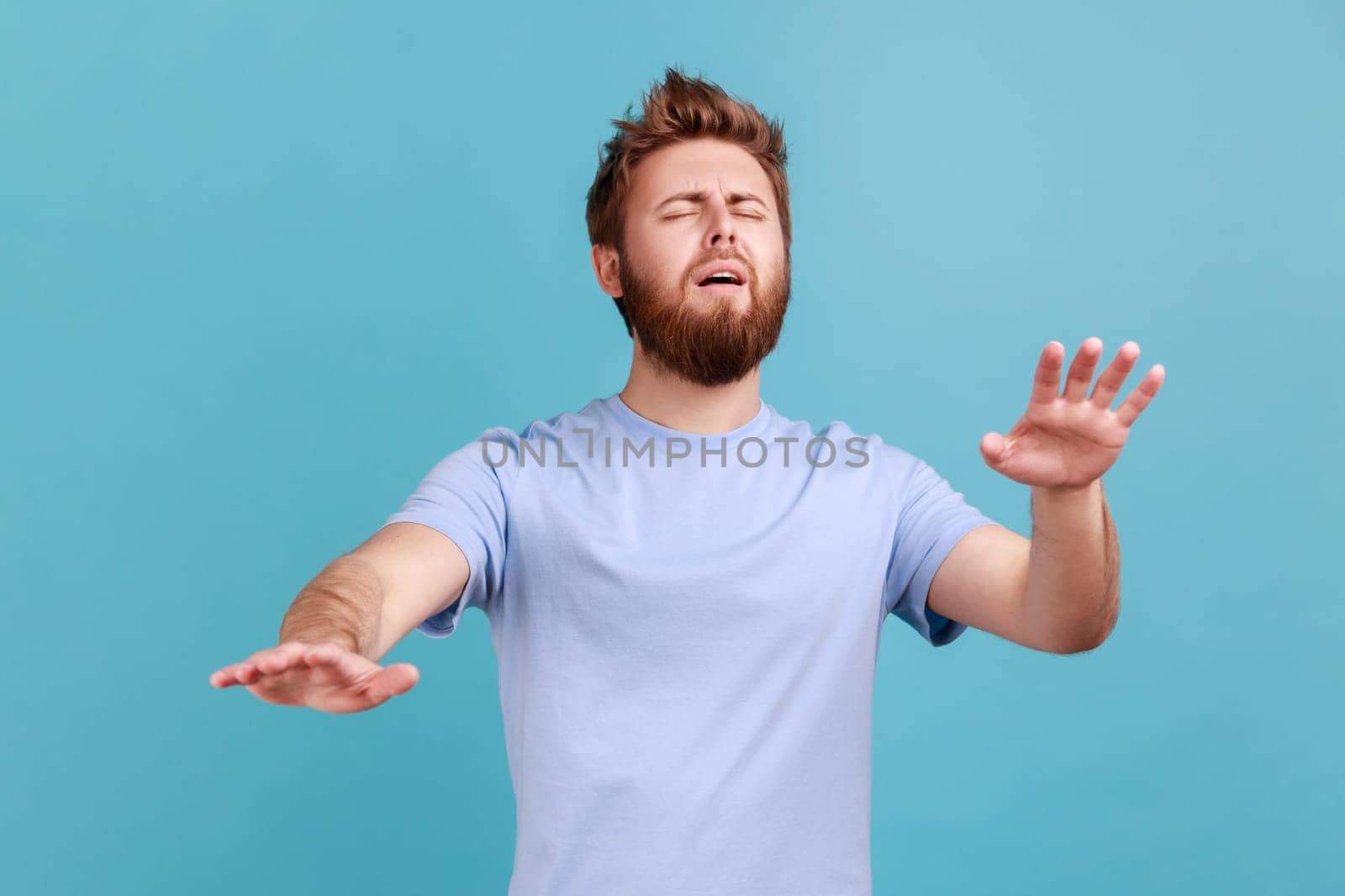 Loneless and blindness. Portrait of sick bearded man standing with closed eyes and try to touching something or find, posing with outstretching hands. Indoor studio shot isolated on blue background.